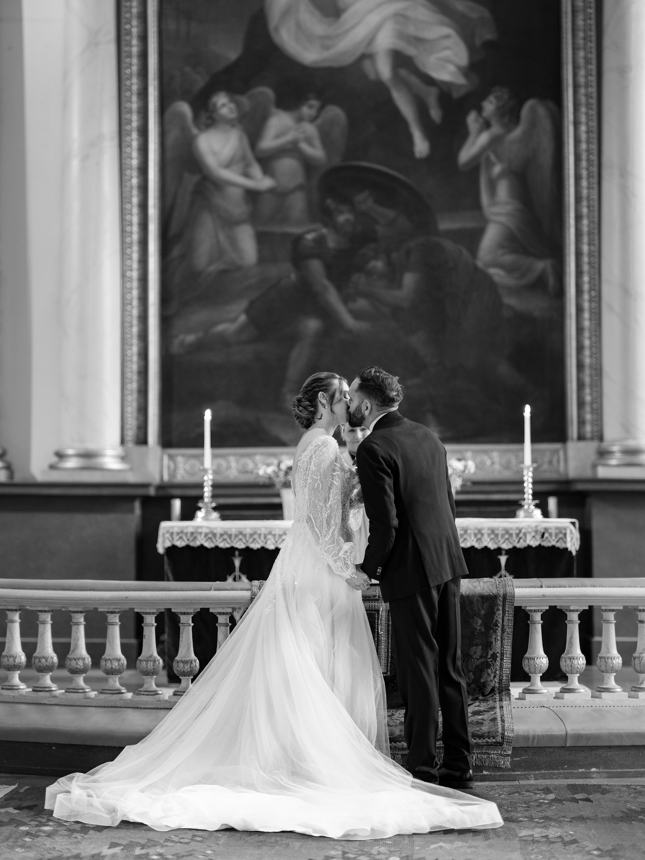 Bride and groom kissing in front of a religious altar with candles and a large painting, photographed in black and white.