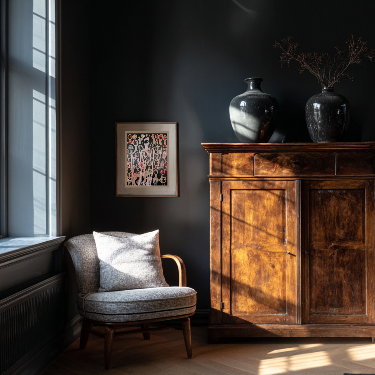 A cozy corner of a room featuring a light-colored upholstered armchair with a matching pillow next to a window, a wooden cabinet with two black vases on top, and a colorful abstract painting on a dark wall.