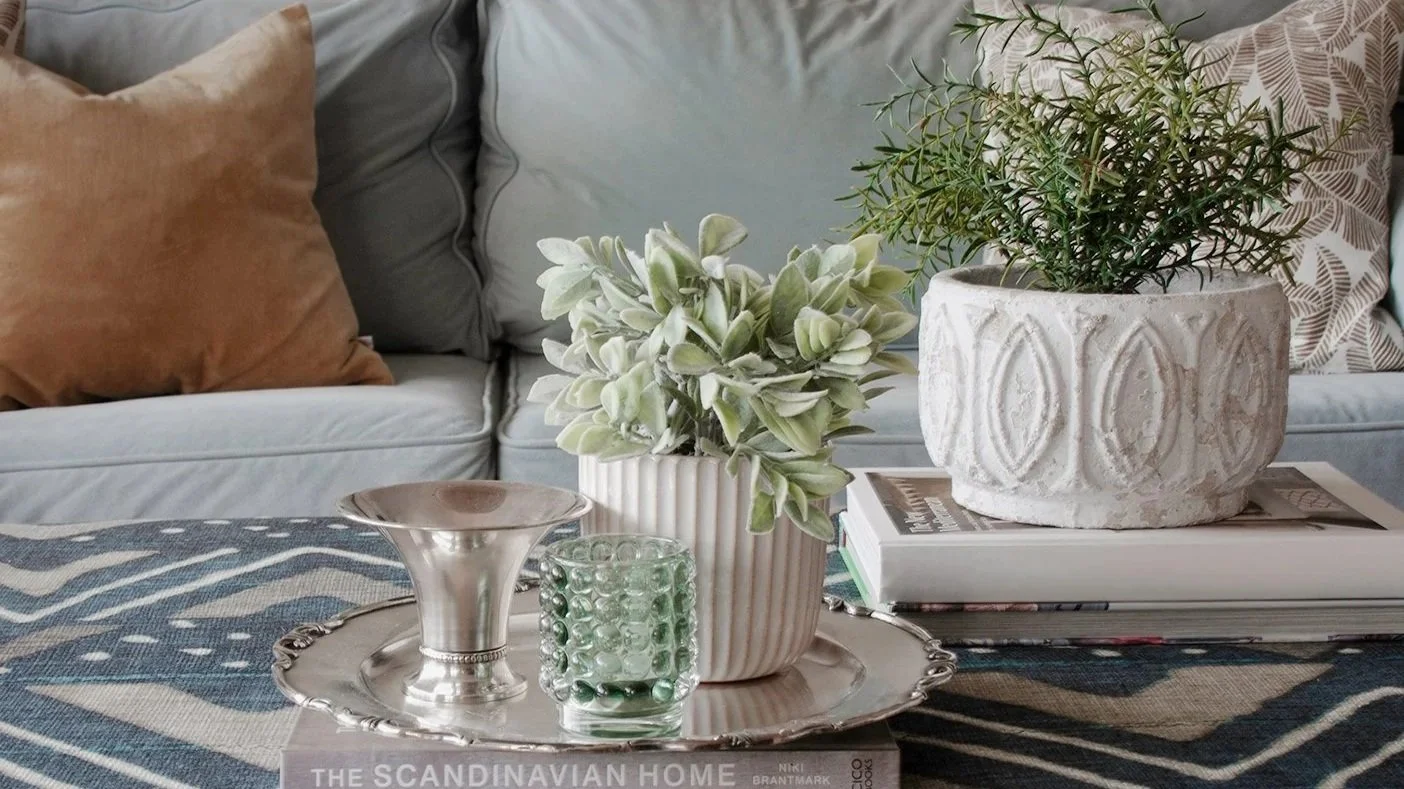 A living room coffee table with decorative items: two potted plants, a small silver tray, a green textured candle, and a stack of magazines. In the background, a light-colored sofa with pillows.