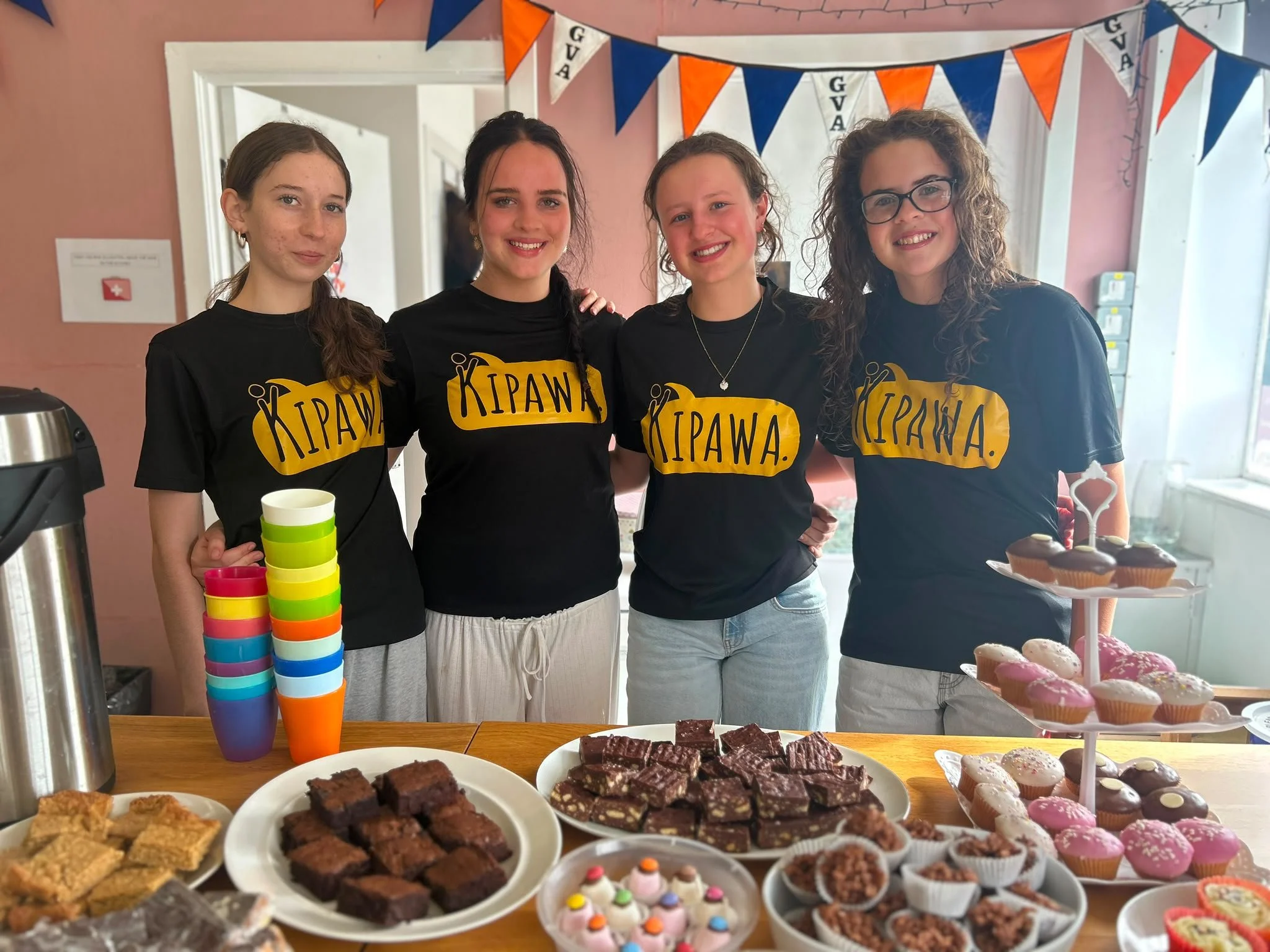 Three young females in black branded Kipawa t-shirts standing in village hall behind table covered in trays of cakes., bakes and flasks of tea and coffee