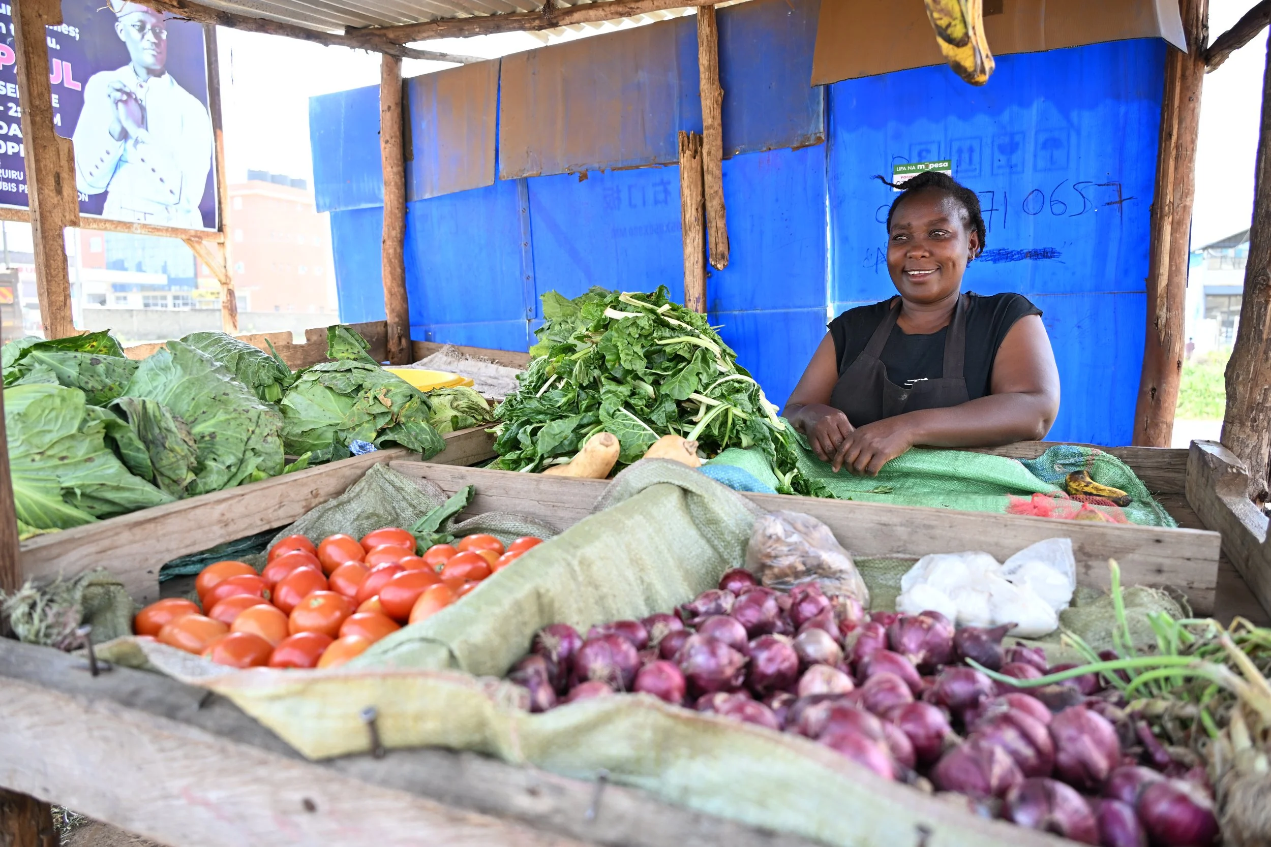 woman standing behind fruit and vegetable stall with red onions, red tomatoes, squash and pile of kale. Bright blue plastic wall in background