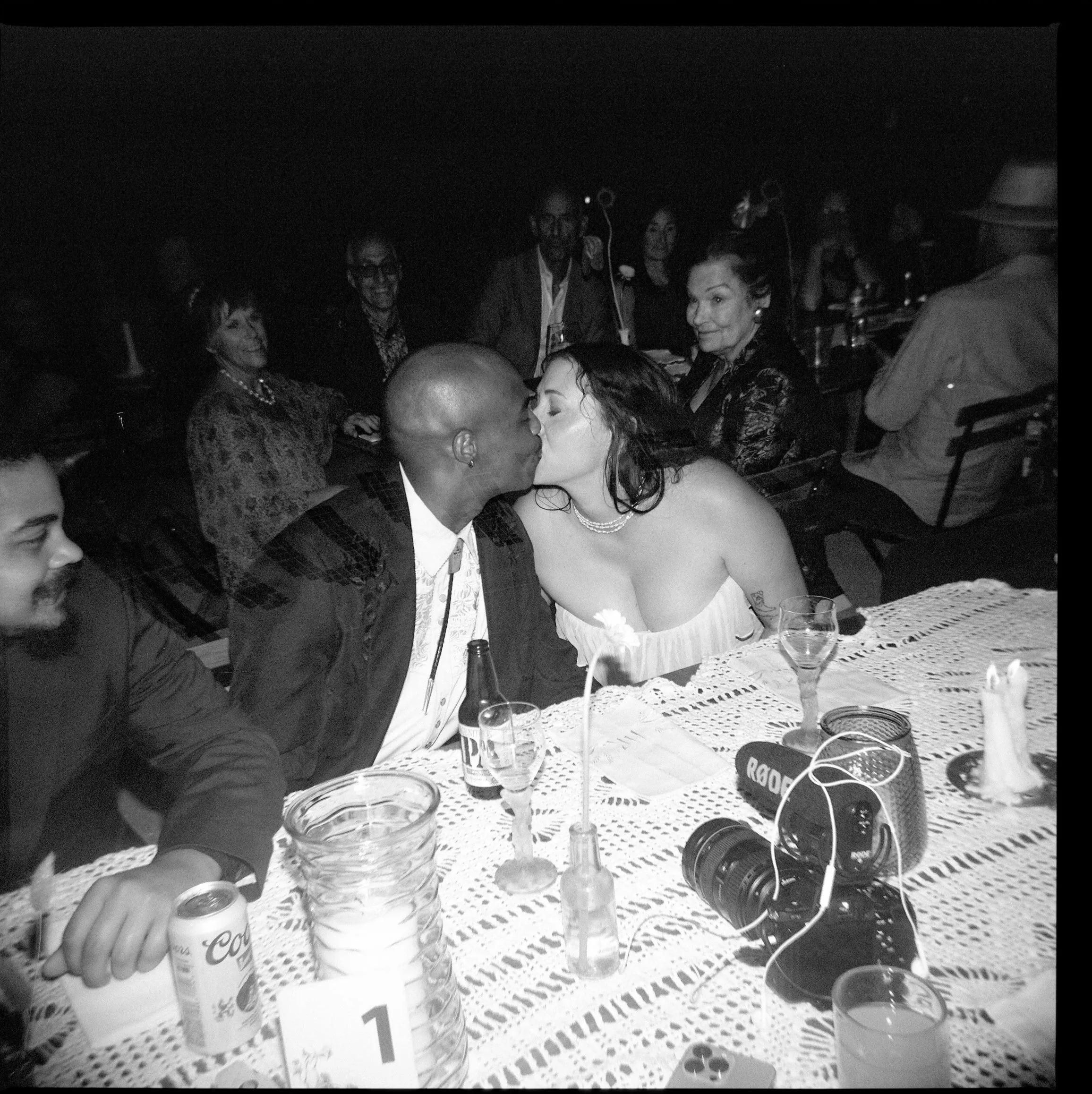 A bride and groom kiss at the dinner table while their guests smile at them
