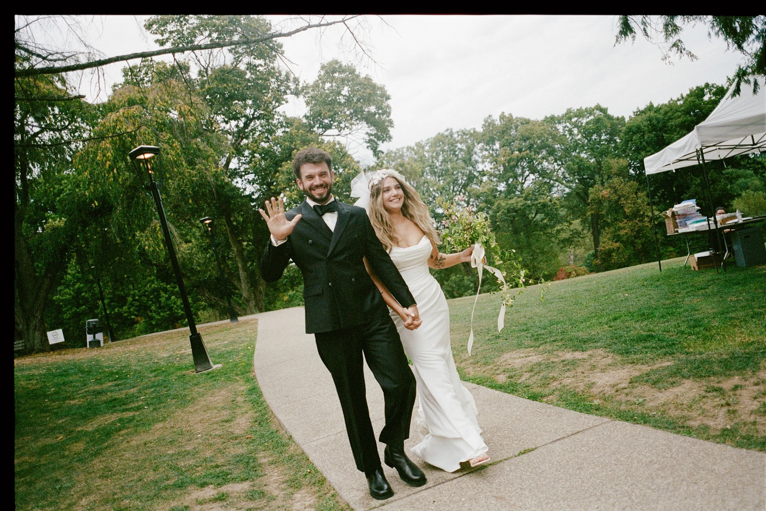 A bride wearing a vintage veil and a groom walk to their reception and wave at the camera