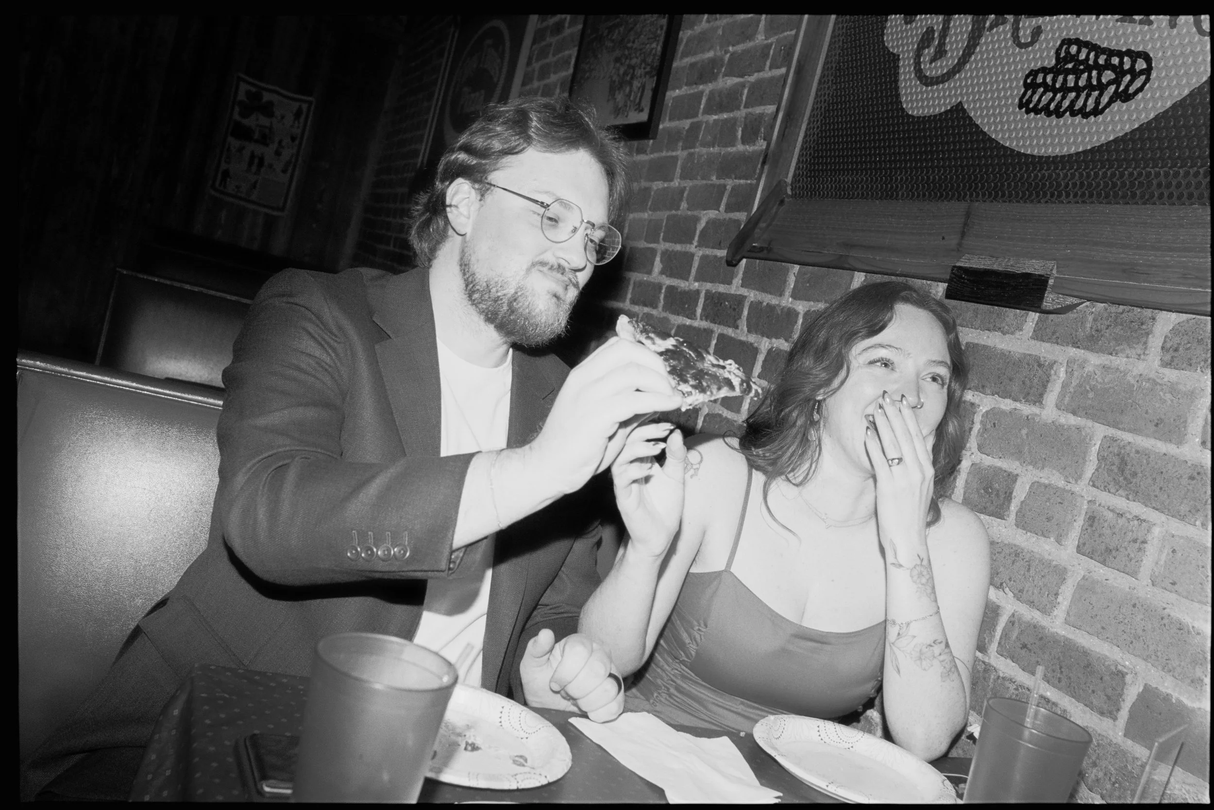 A man with glasses and a beard is feeding a slice of pizza to a woman with wavy hair, who is laughing and covering her mouth with her hand. They are sitting at a table in a restaurant with brick walls.