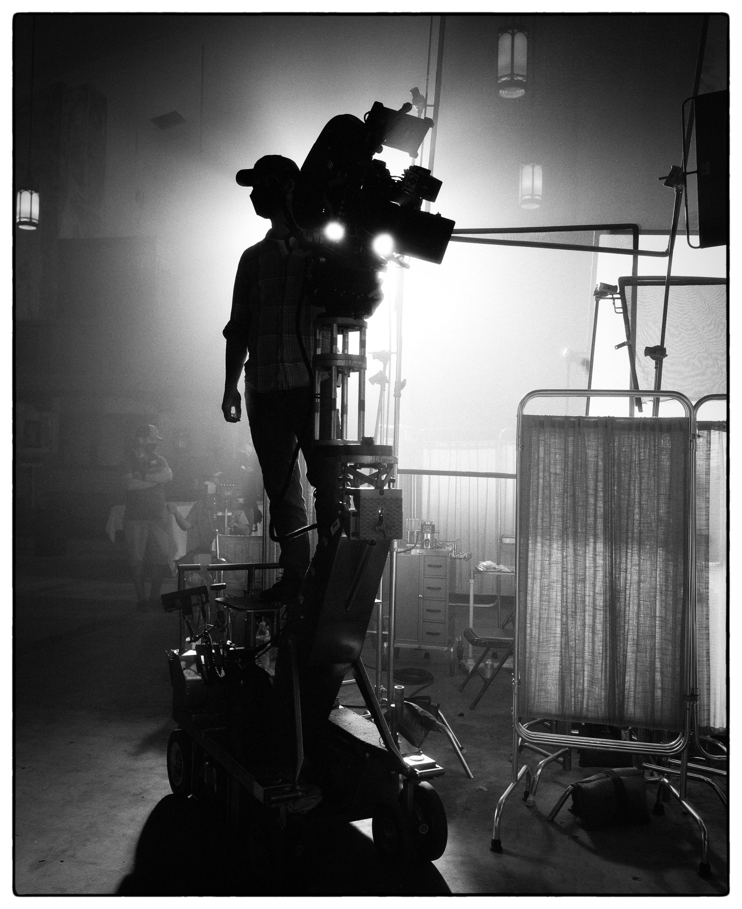 A man standing on a camera dolly with a Arri film camera mounted on it. He is in silhouette.