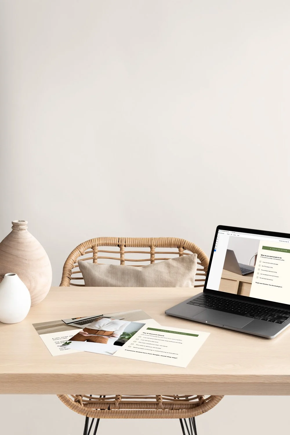A minimalist workspace with a wooden table, two ceramic vases, a rattan chair with a cushion, and a laptop displaying a form, set against a plain off-white wall.