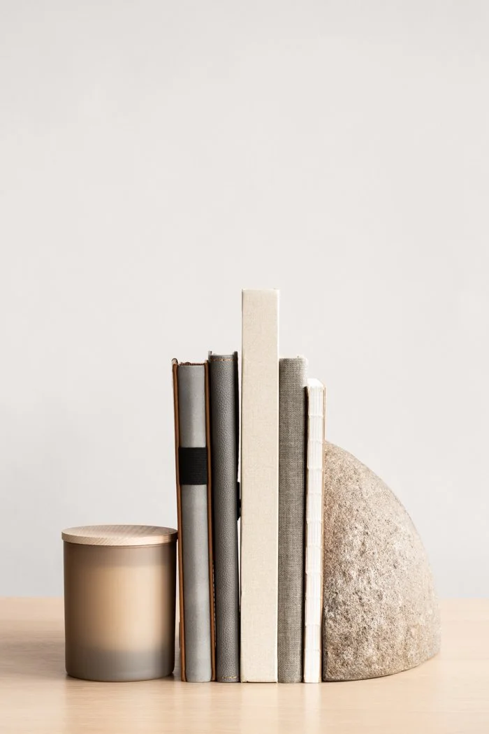 A collection of six books of varying sizes with neutral-colored covers, arranged upright on a light wooden surface, with a rounded stone object next to the rightmost book, and a candle with a frosted glass holder and wooden lid on the left.