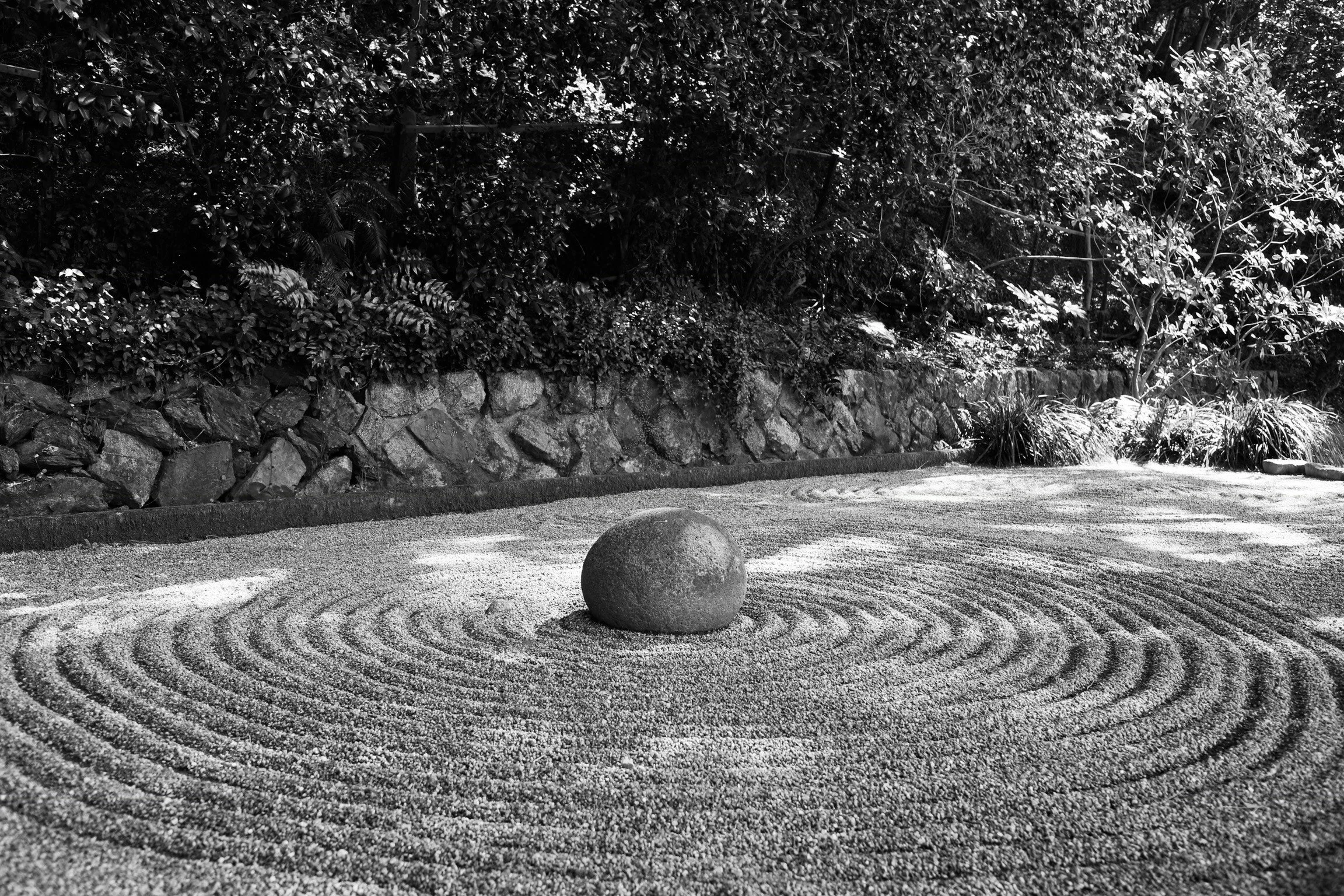 A traditional Japanese Zen garden with a large round stone at the center, surrounded by carefully raked gravel in concentric circles, and a stone wall with lush foliage in the background.