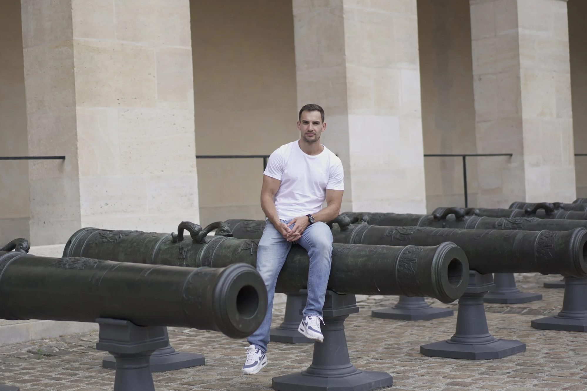 Un homme assis sur une vieille pièce d'artillerie en extérieur, dans un lieu historique avec des colonnes en pierre.