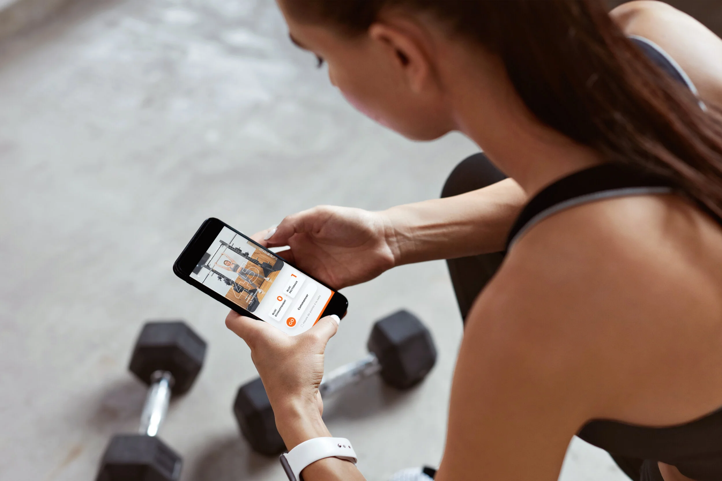 Une femme faisant des exercices de musculation avec des haltères, regardant une application de fitness sur son smartphone pour se former, portant un bracelet connecté.