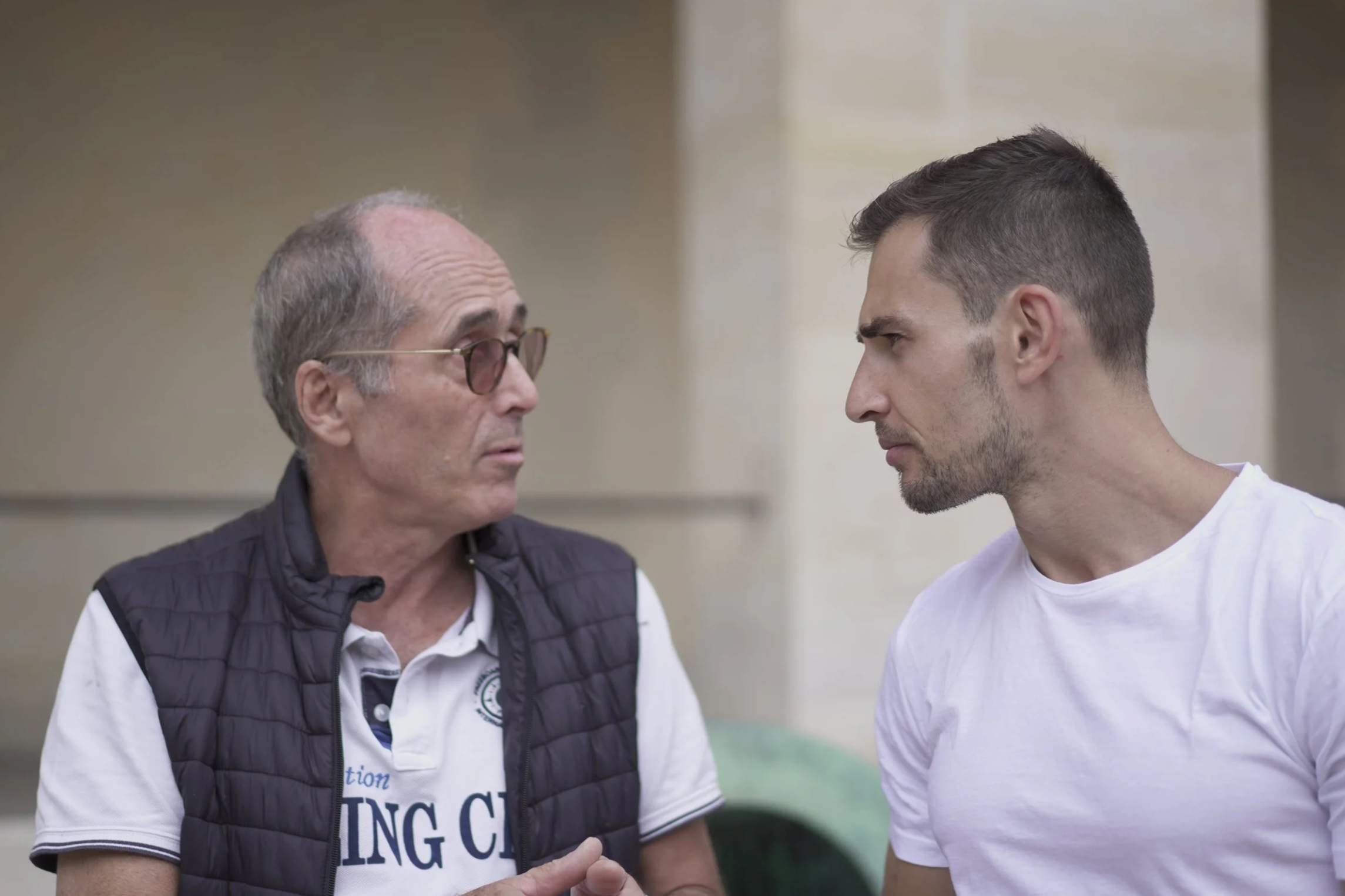 Deux hommes en conversation, l'un âgé portant des lunettes et un gilet, l'autre plus jeune en t-shirt blanc, se regardant avec sérieux dans un lieu intérieur.