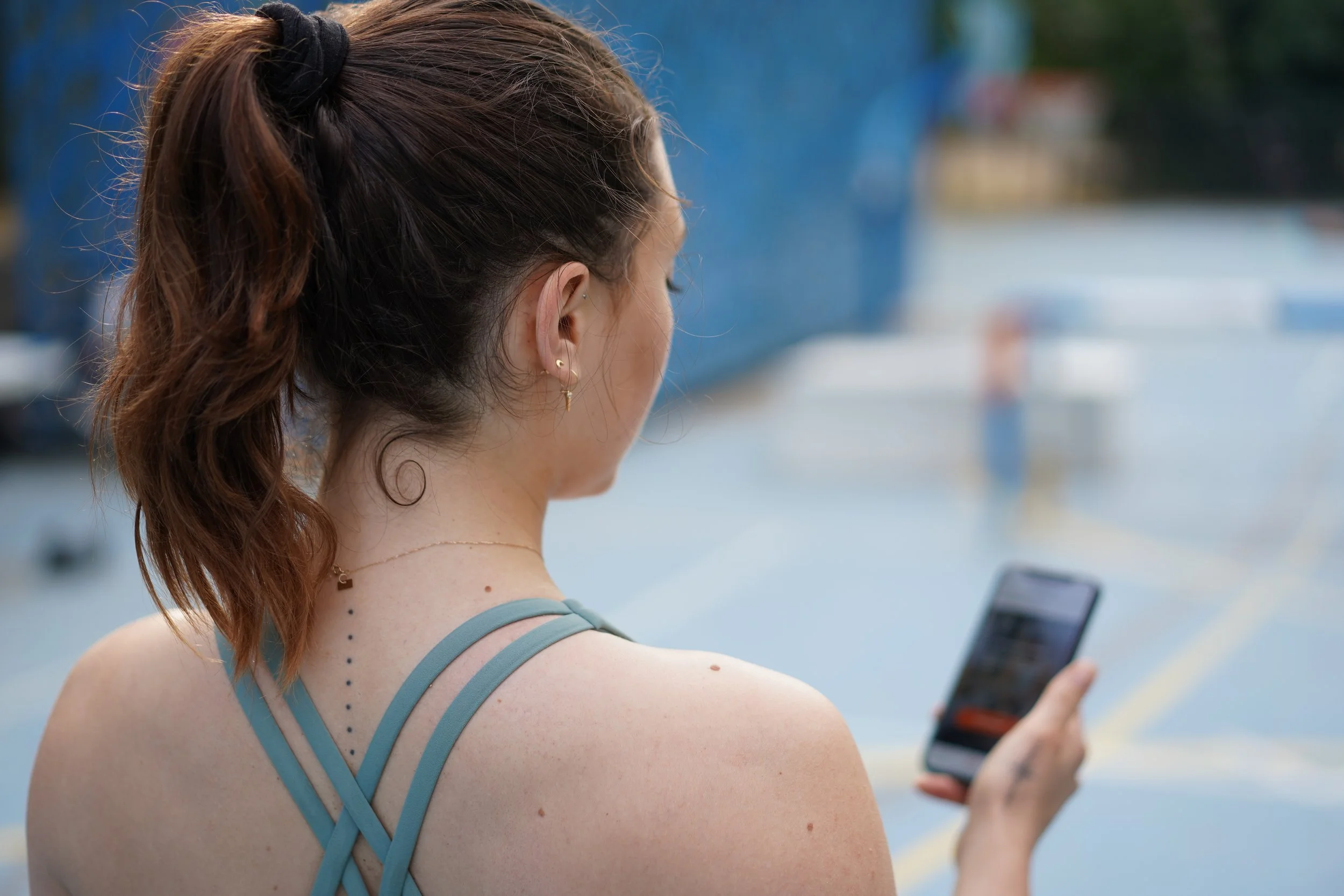 Une femme vue de dos, avec des cheveux bruns attachés en queue de cheval, portant un haut bleu avec des bretelles croisées, regardant son téléphone dans un espace extérieur.