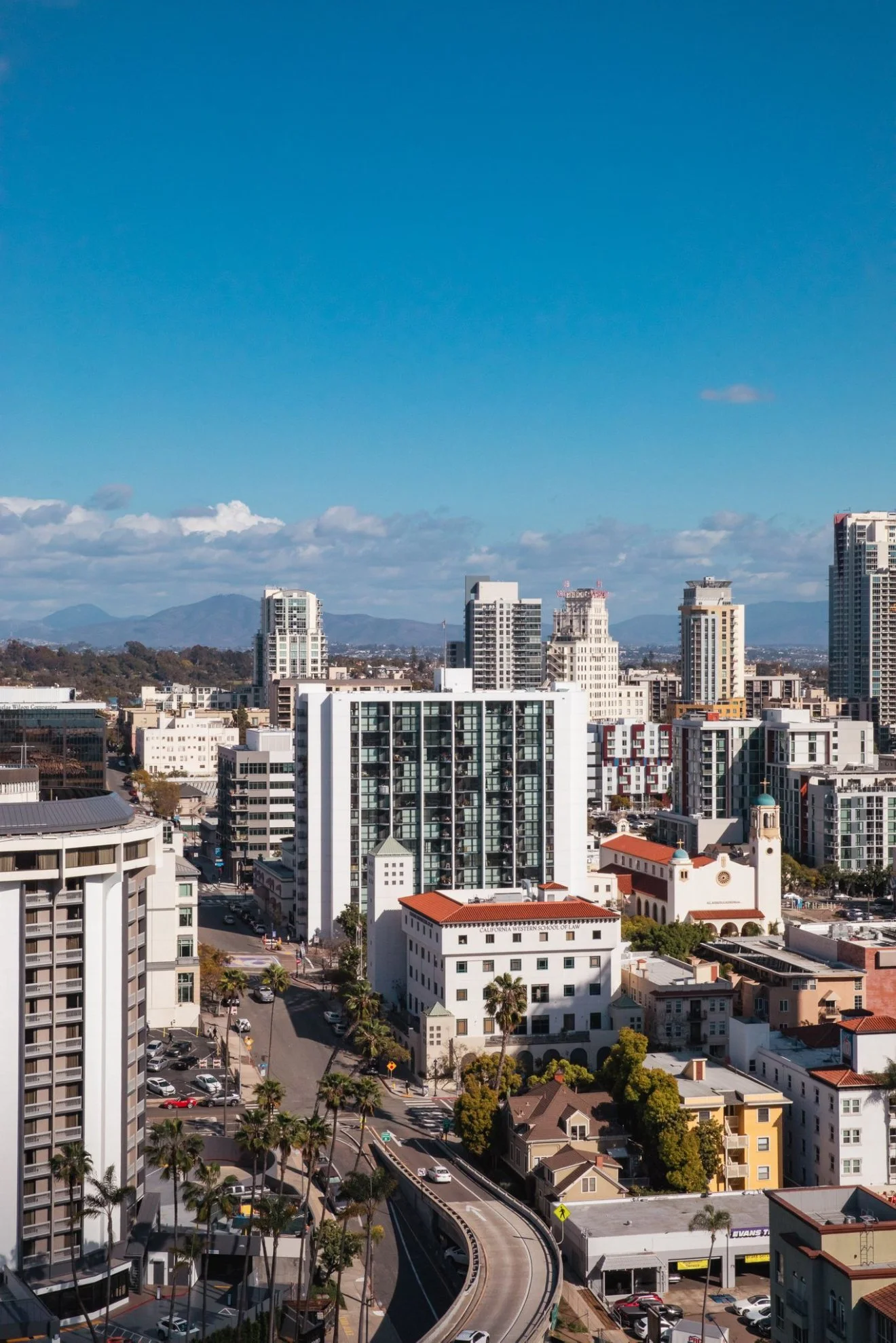 Cityscape of downtown with tall buildings, a church with a steeple, palm trees, and residential structures under a partly cloudy blue sky.