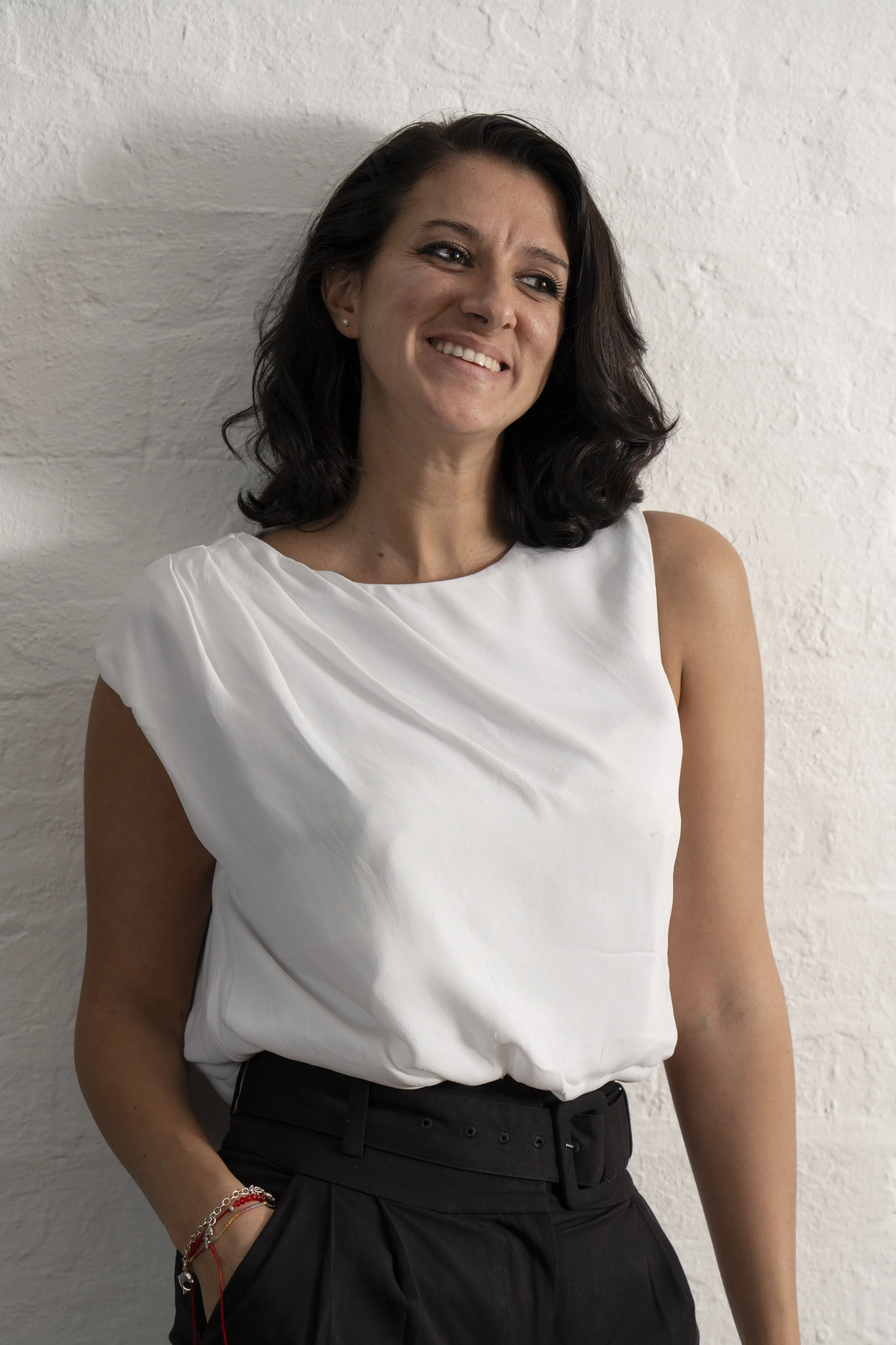 Female with dark hair and white top poses for a relaxed corporate portrait by leaning against a white wall