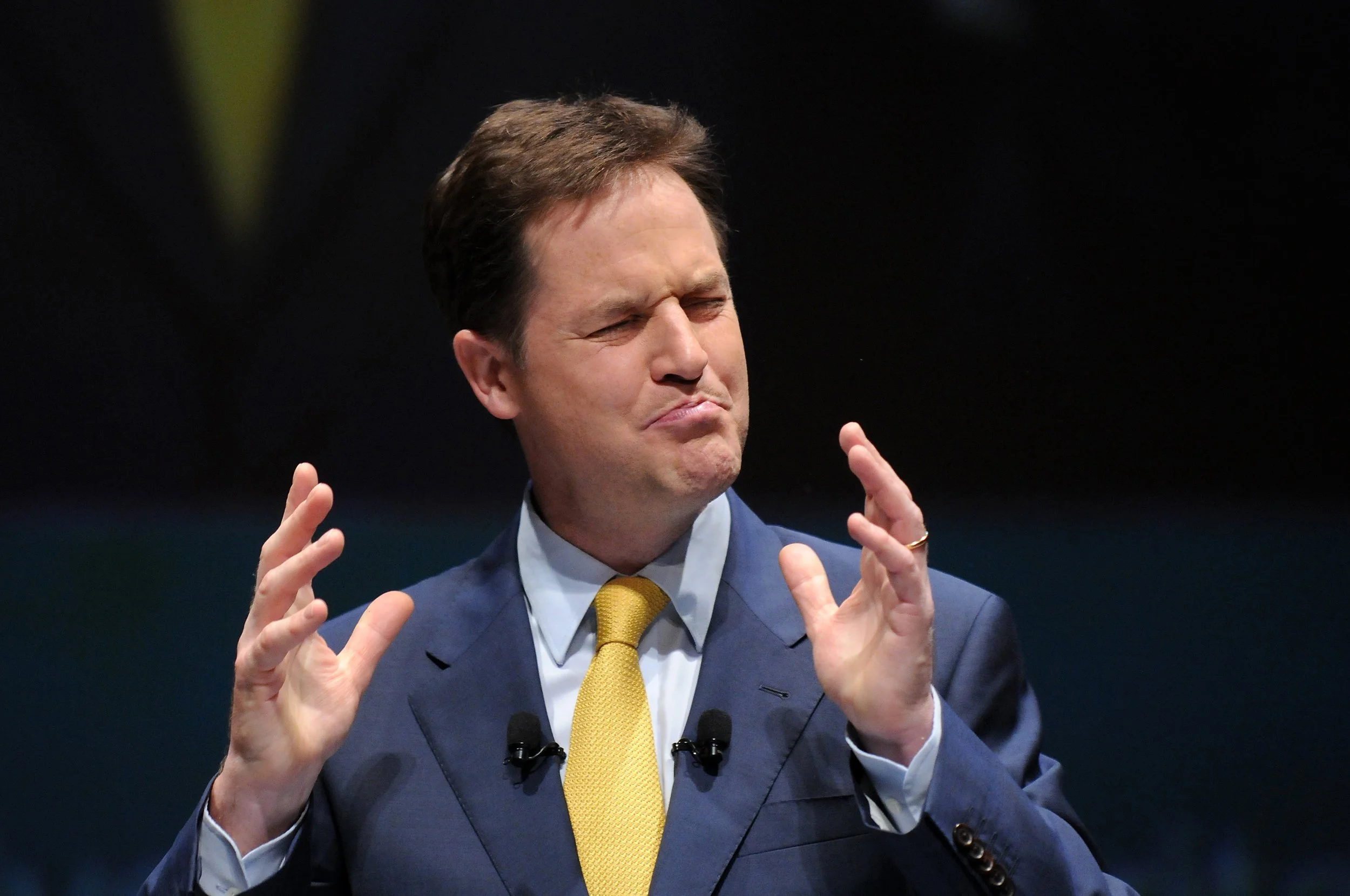 A man wearing a blue suit, light blue shirt, and yellow tie with a pained or confused facial expression, eyes closed, and hands raised near his face.