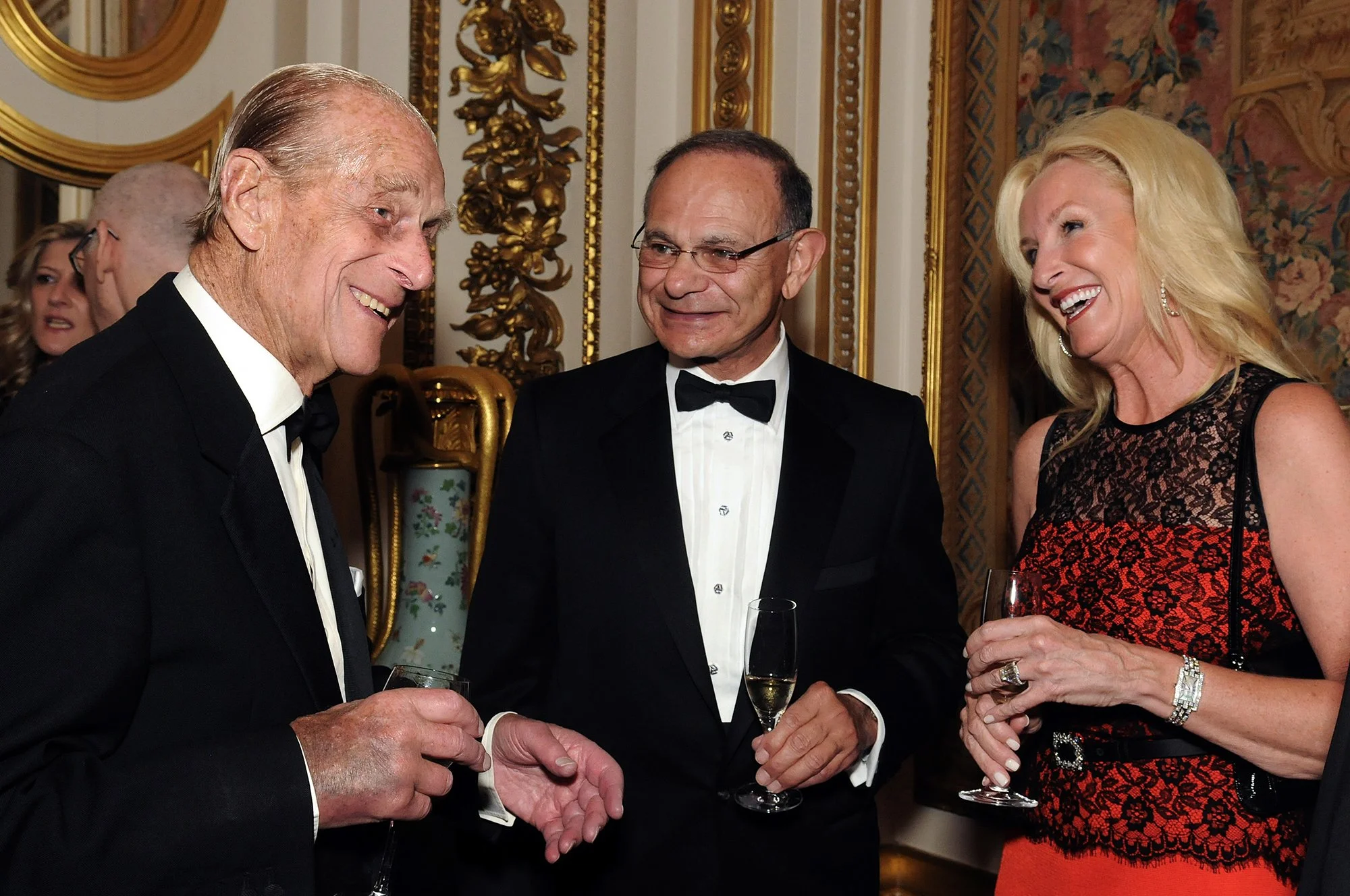 Three well-dressed people, two men in tuxedos and a woman in a sleeveless black and red dress, are smiling and talking at a formal event, holding champagne glasses.