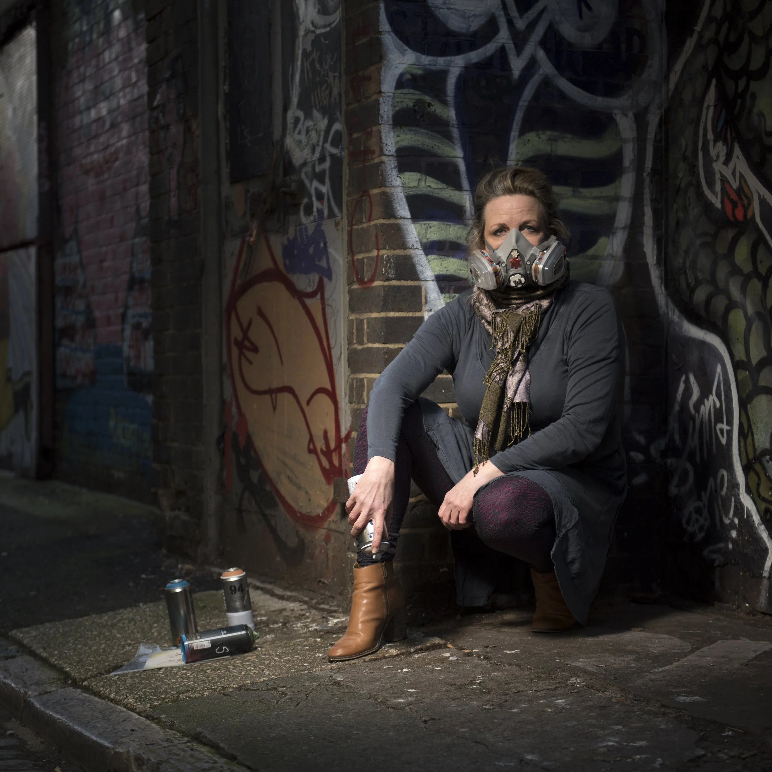 Portrait of a female graffiti artist crouching on the pavement next to a graffiti covered wall whilst wearing a mask