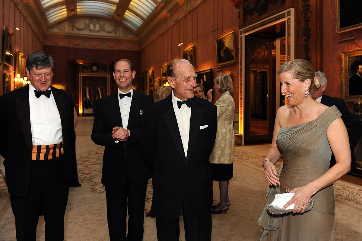 Elegant women and men in tuxedos and evening gowns talking and smiling at a formal event in a luxurious room with ornate decor and framed paintings.