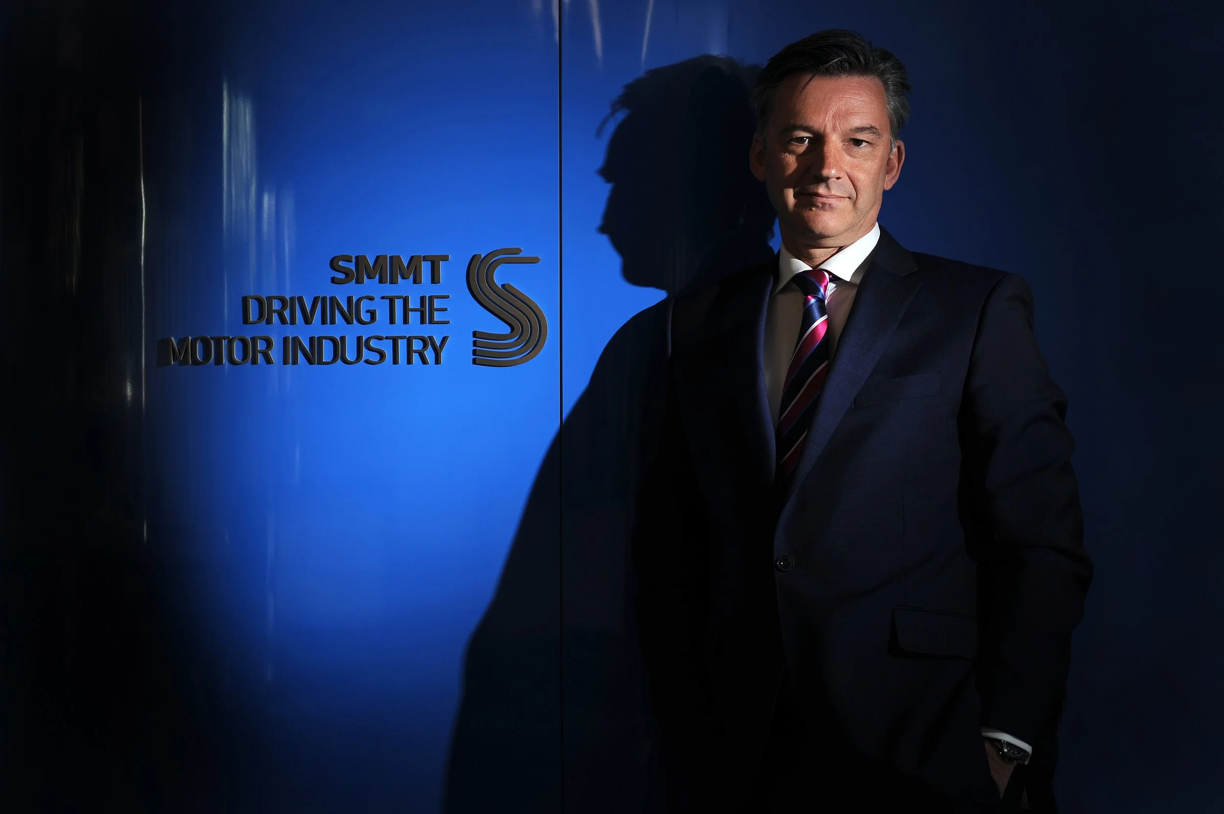 A man in a suit standing in front of a blue wall with the logo and text 'SMMT Driving the Motor Industry'.