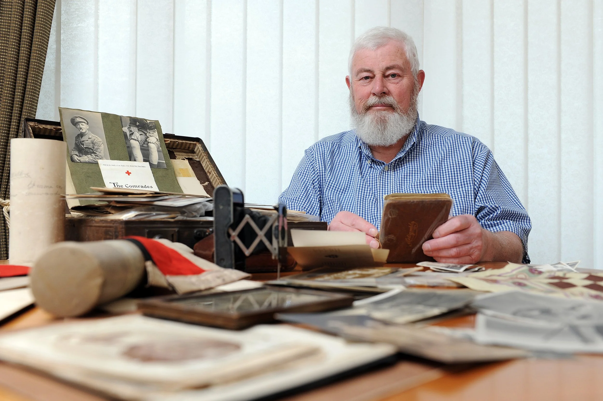 An elderly man with white hair and a beard, sitting at a desk cluttered with photographs, papers, and objects, holding a book and looking at the camera. Behind him is a window with closed blinds.