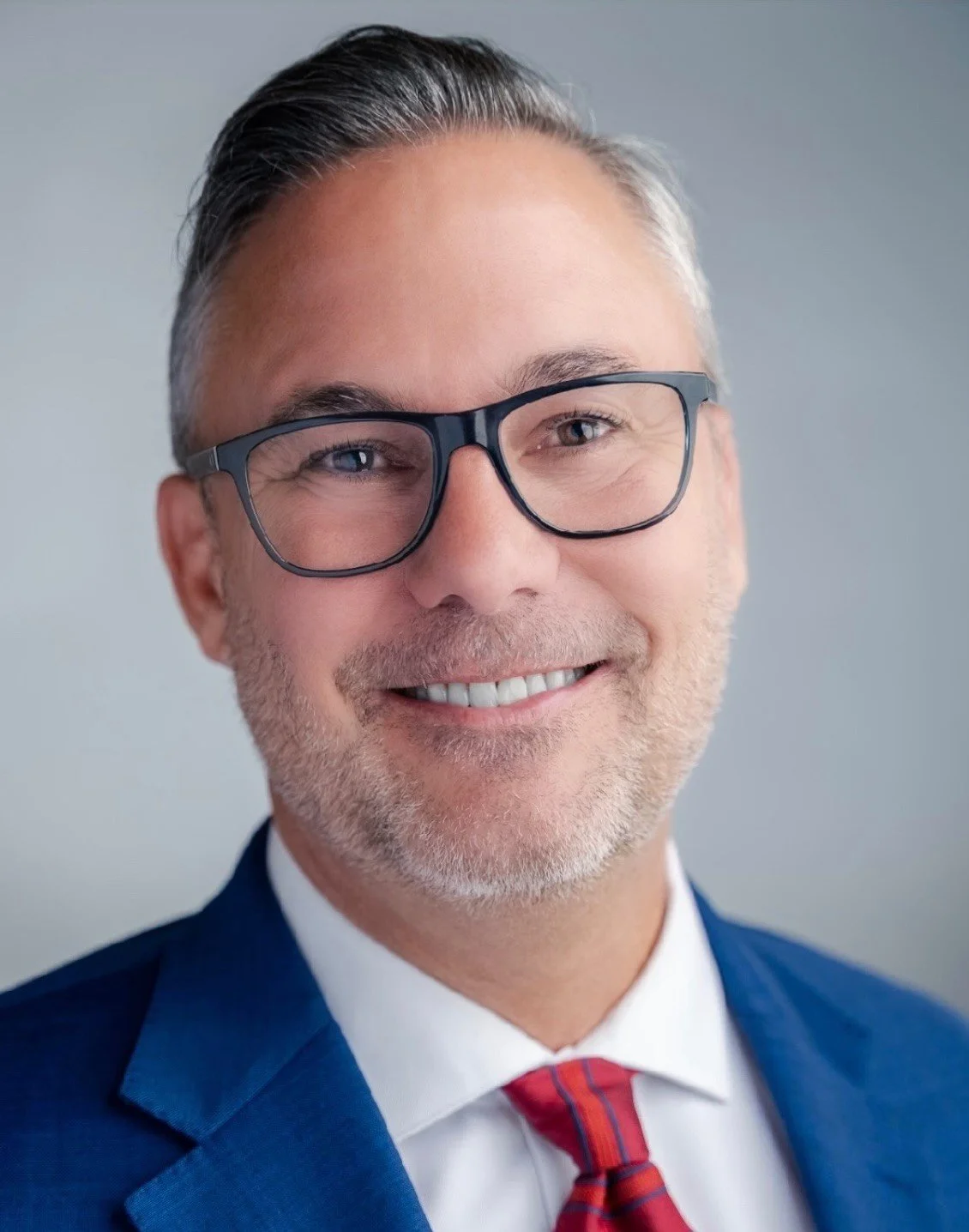 A smiling middle-aged man with gray hair and glasses, dressed in a blue suit, white shirt, and red striped tie, posing against a neutral background.