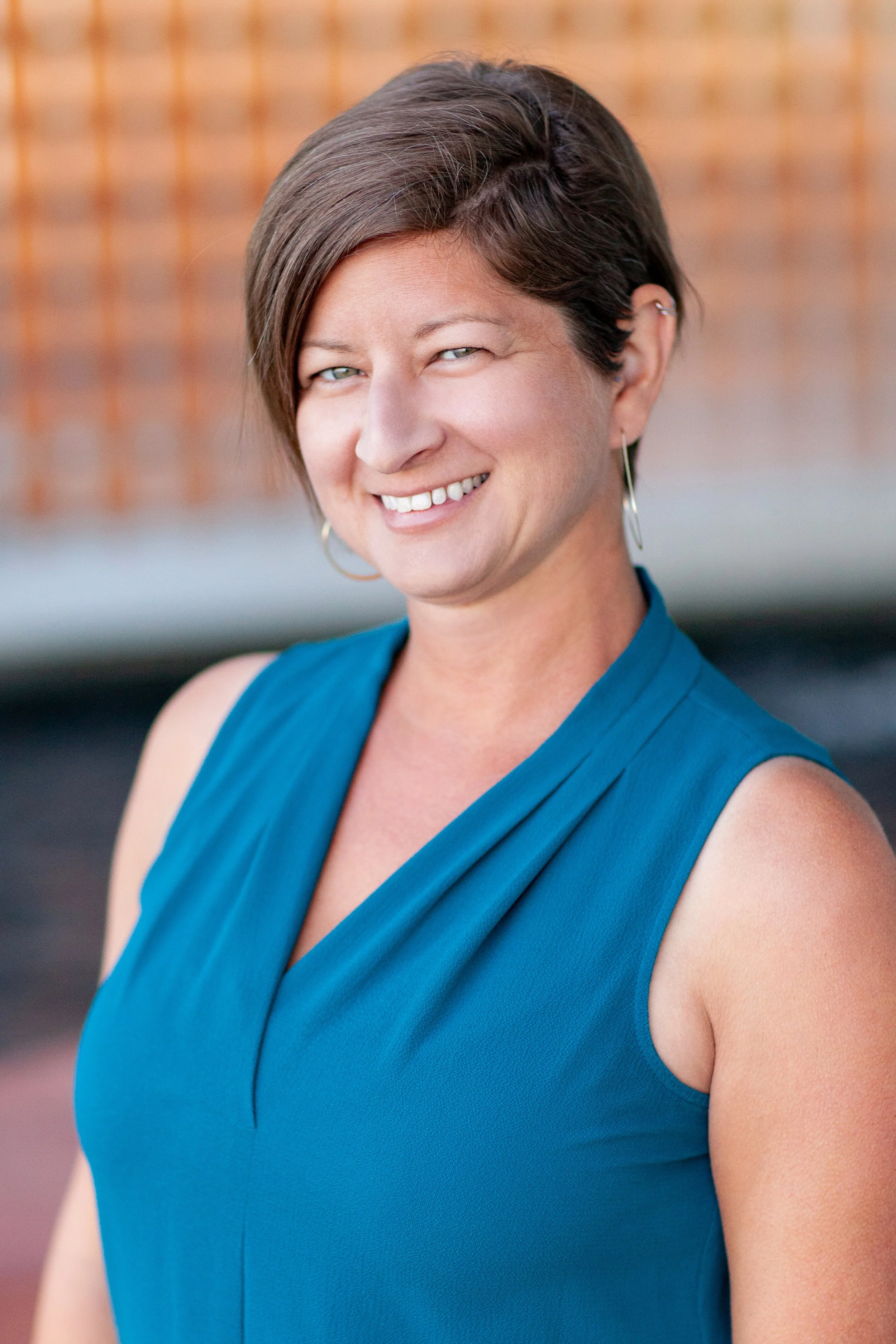 A smiling woman with short brown hair wearing a teal sleeveless top and hoop earrings, standing outdoors with a blurred brick wall background.