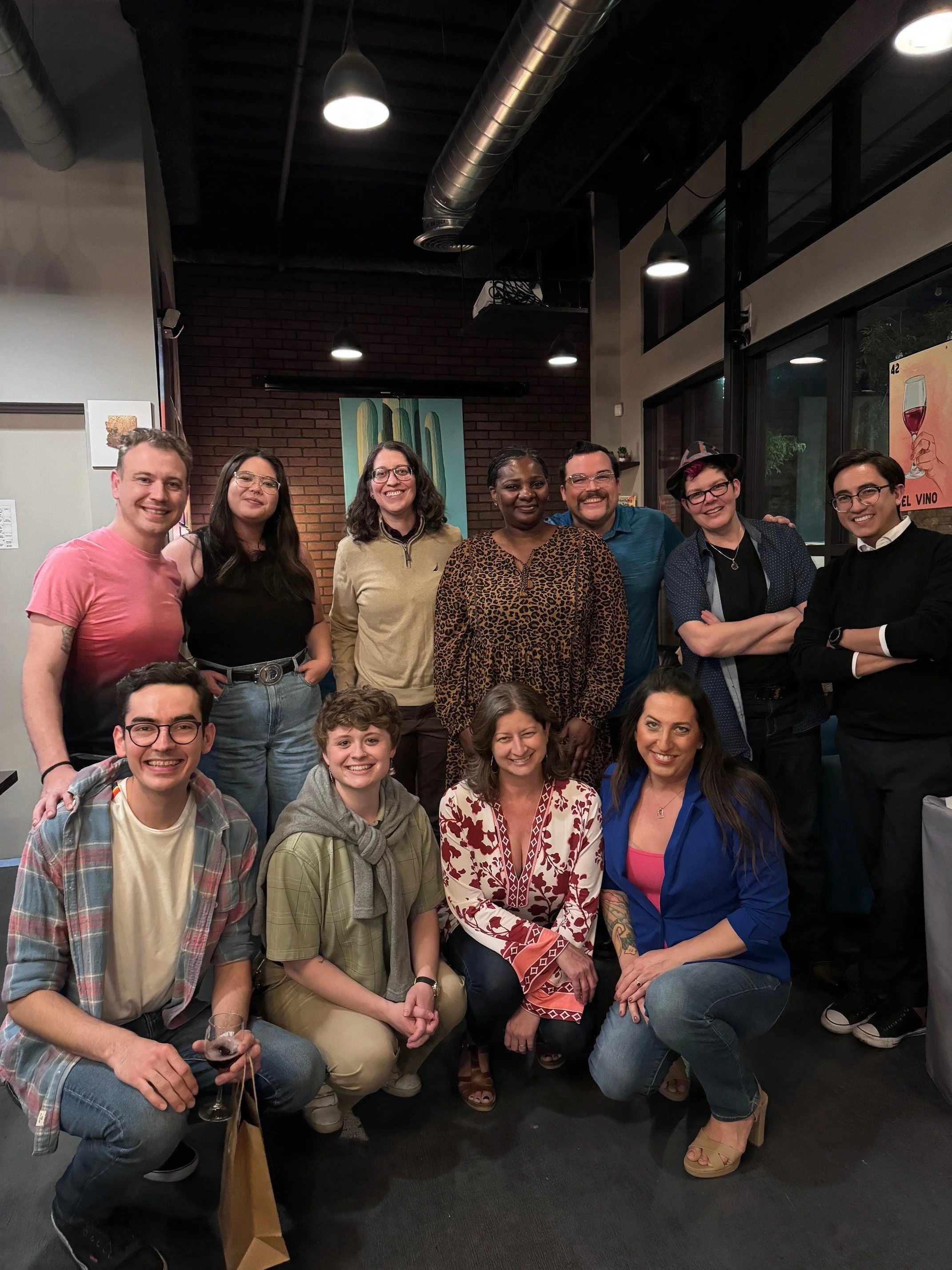 A diverse group of eleven people, including men and women of various ethnicities, gathered together in a restaurant or cafe. They are smiling and posing for the photo, with some standing and others squatting in front. The background features a brick wall, artwork, and large windows.