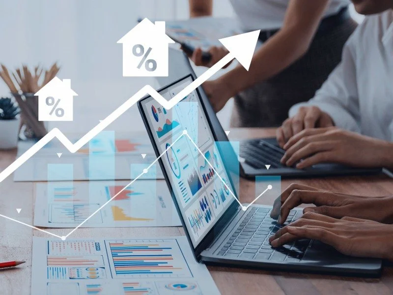 Two people reviewing real estate market charts on a laptop and printed reports, with a rising arrow and house icons marked with percent signs.