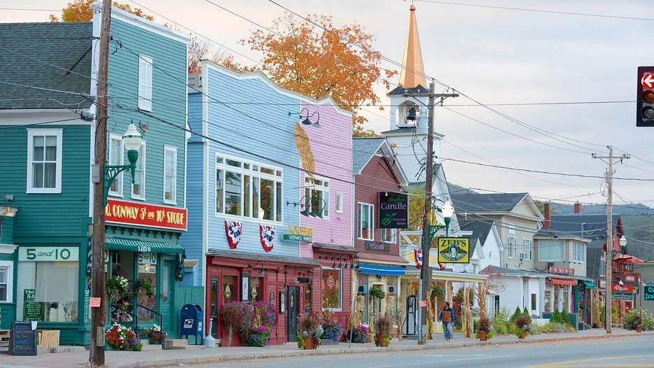 Colorful buildings along a street with a church steeple in the background, autumn trees, and a few pedestrians.