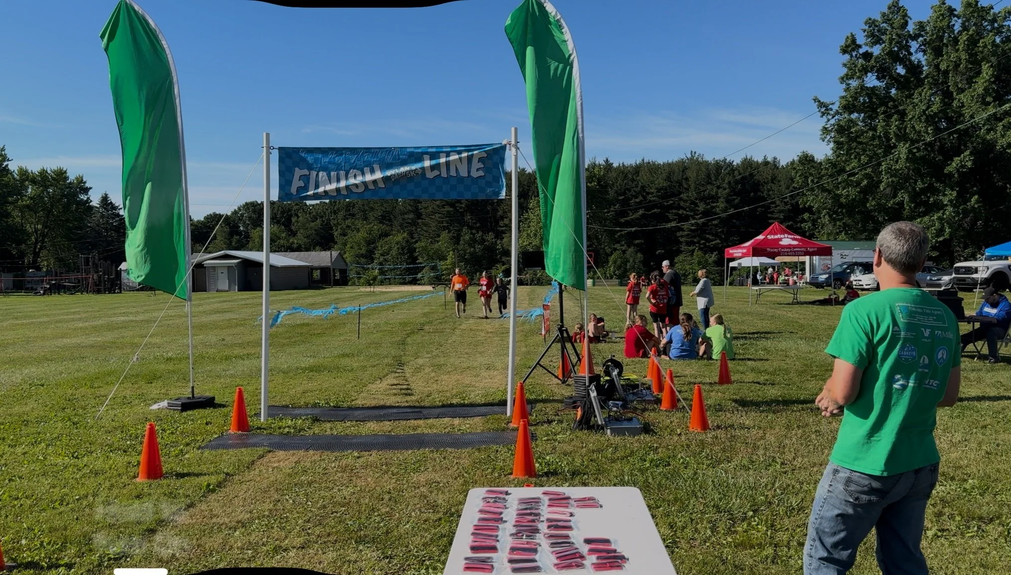 Outdoor race finish line with runners approaching, surrounded by green flags and orange cones, people watching nearby, red tent in background, sunny day.