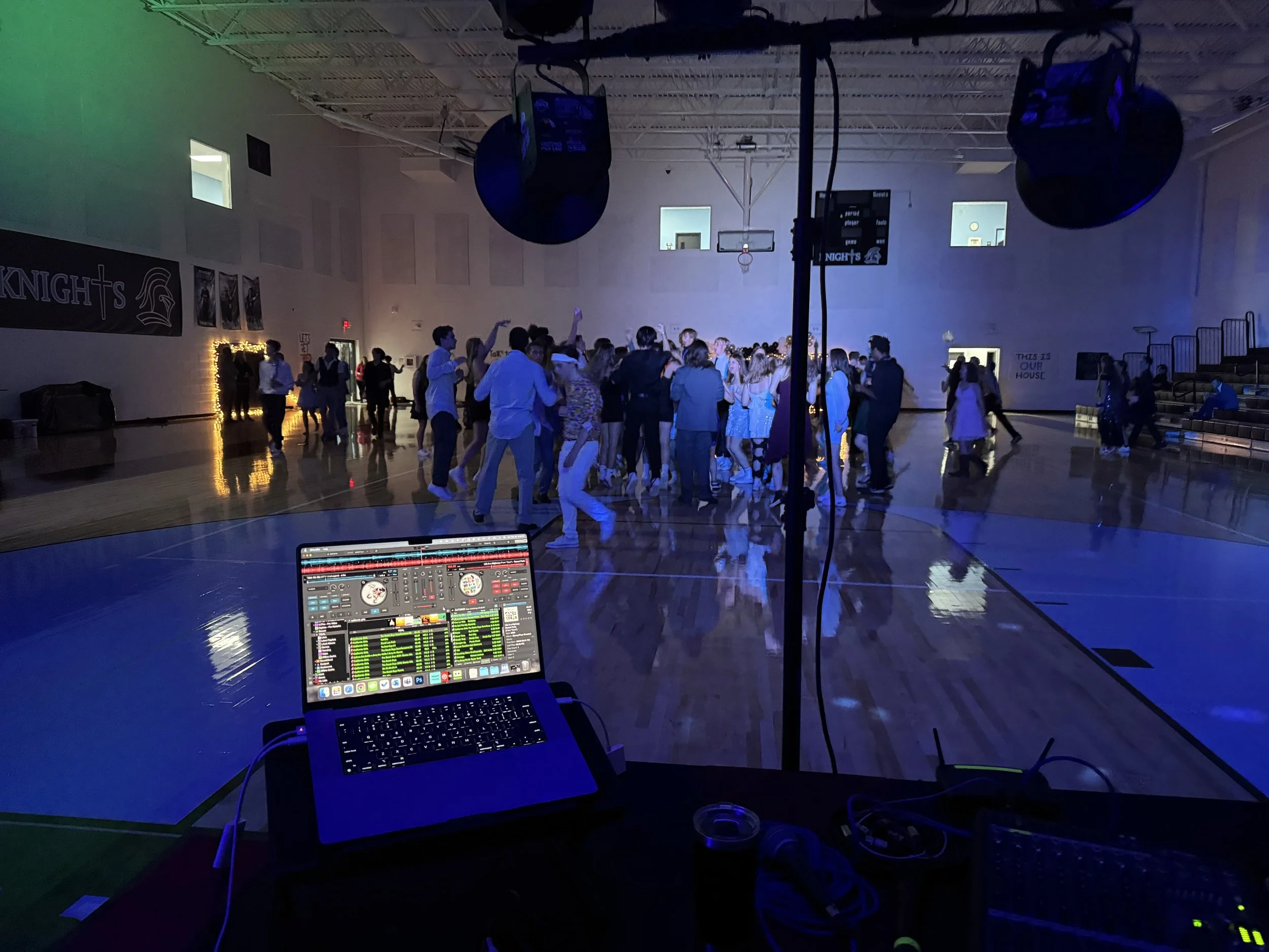 A dimly lit gym with people dancing during a party, viewed from a DJ booth with a laptop and audio equipment.