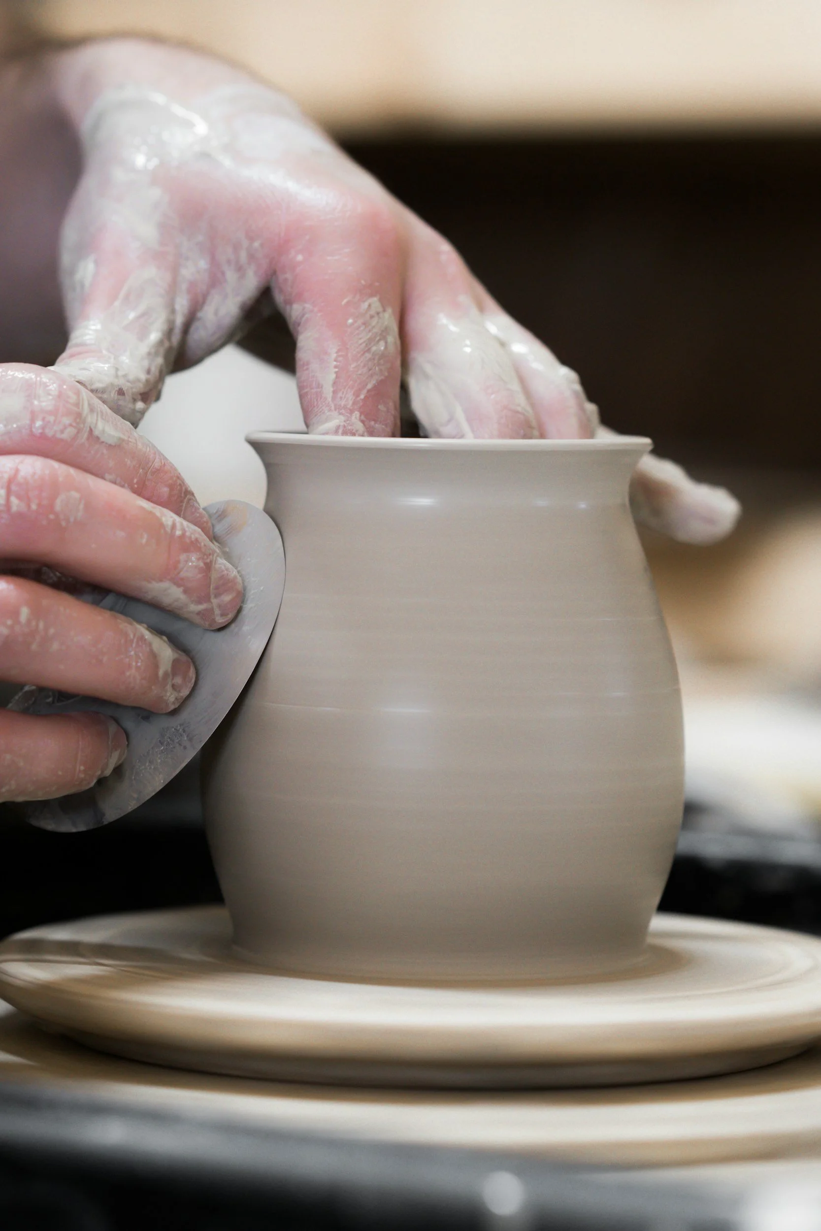 Close-up of a person's hands shaping a ceramic vase on a pottery wheel, wearing gloves and covered in clay.