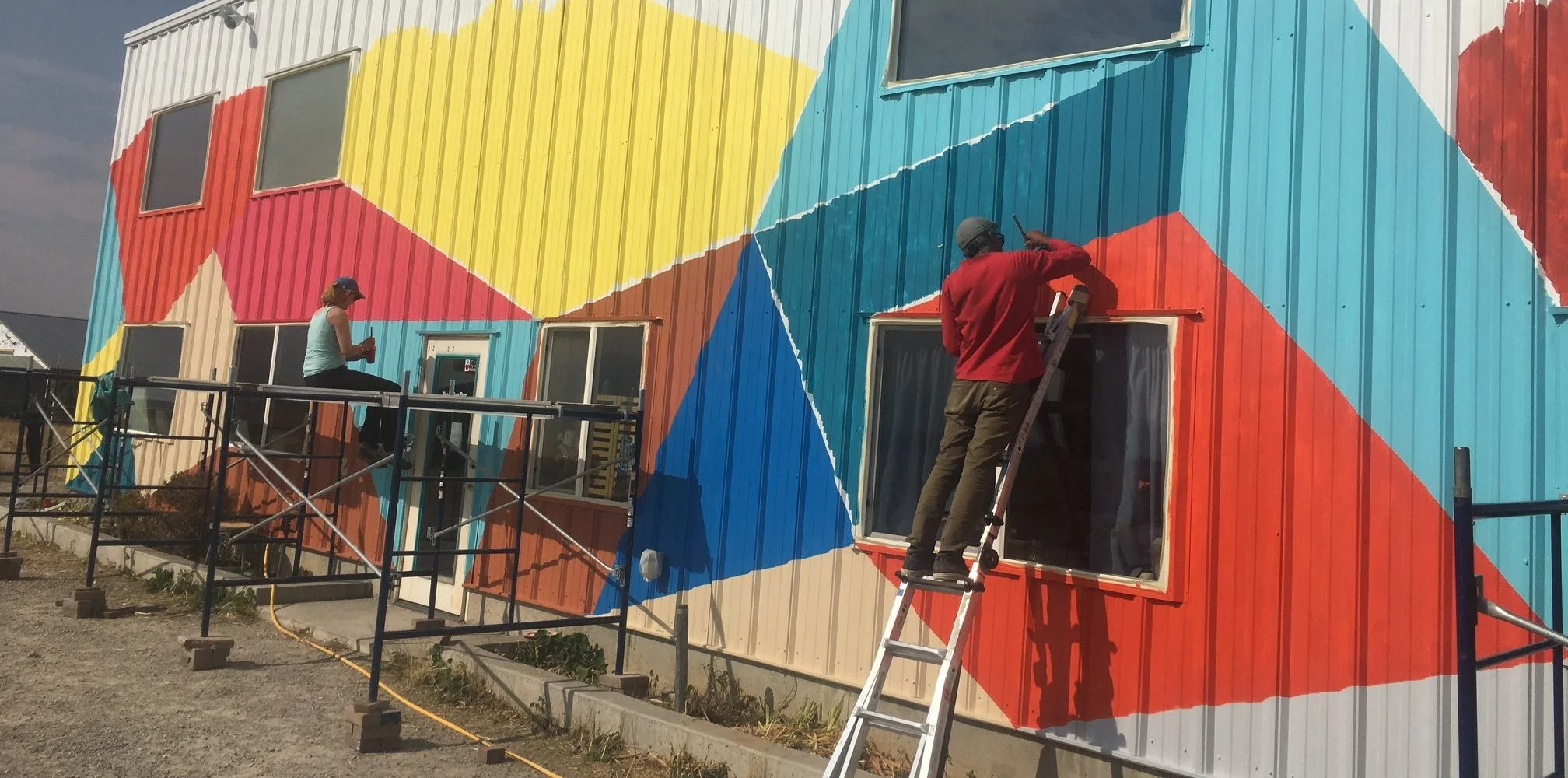 Two people painting a multicolored geometric mural on the exterior wall of a building, one sitting on scaffolding and the other standing on a ladder.