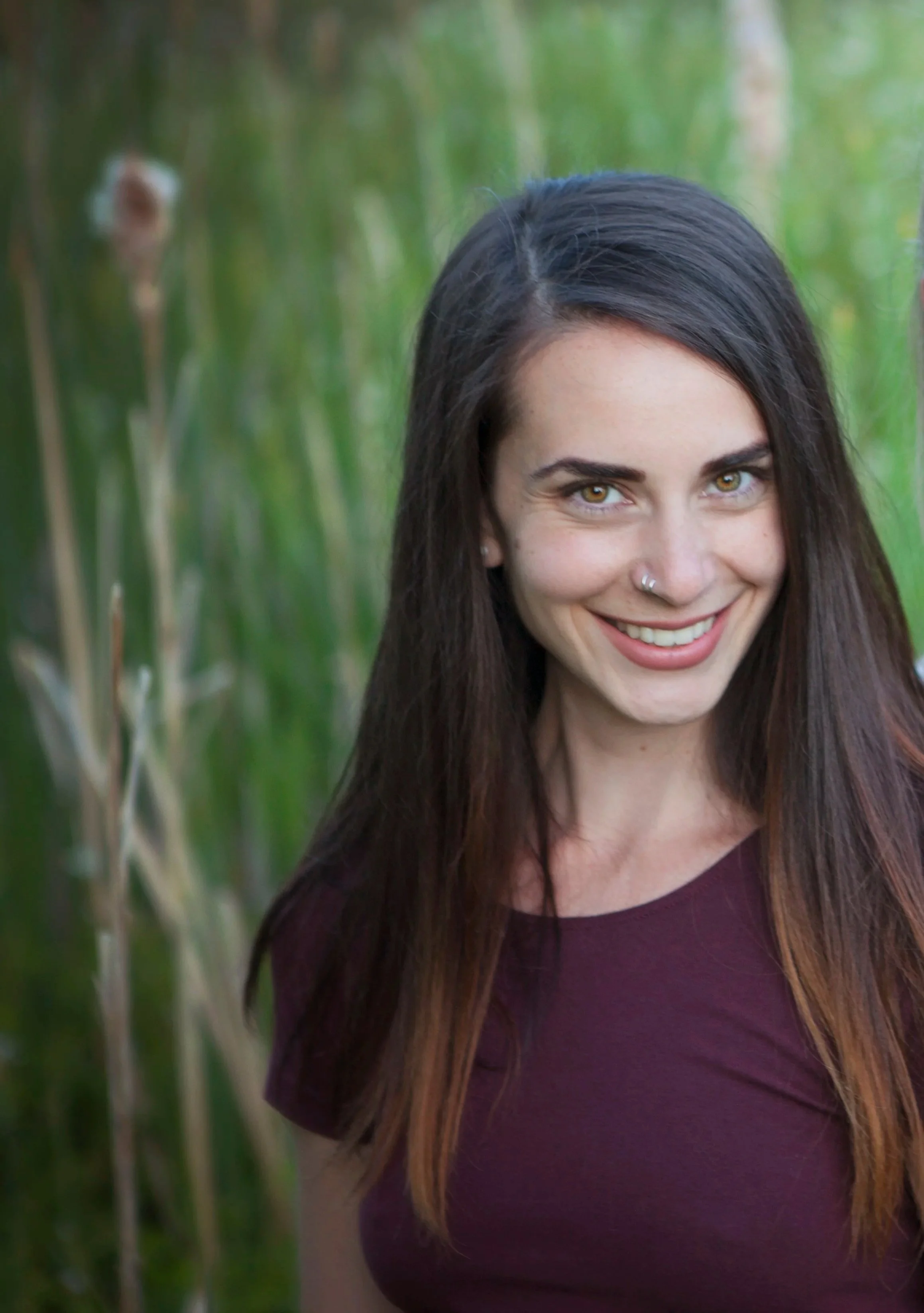 A woman with long dark hair, hazel eyes, and a nose piercing smiles at the camera outdoors with green foliage in the background.