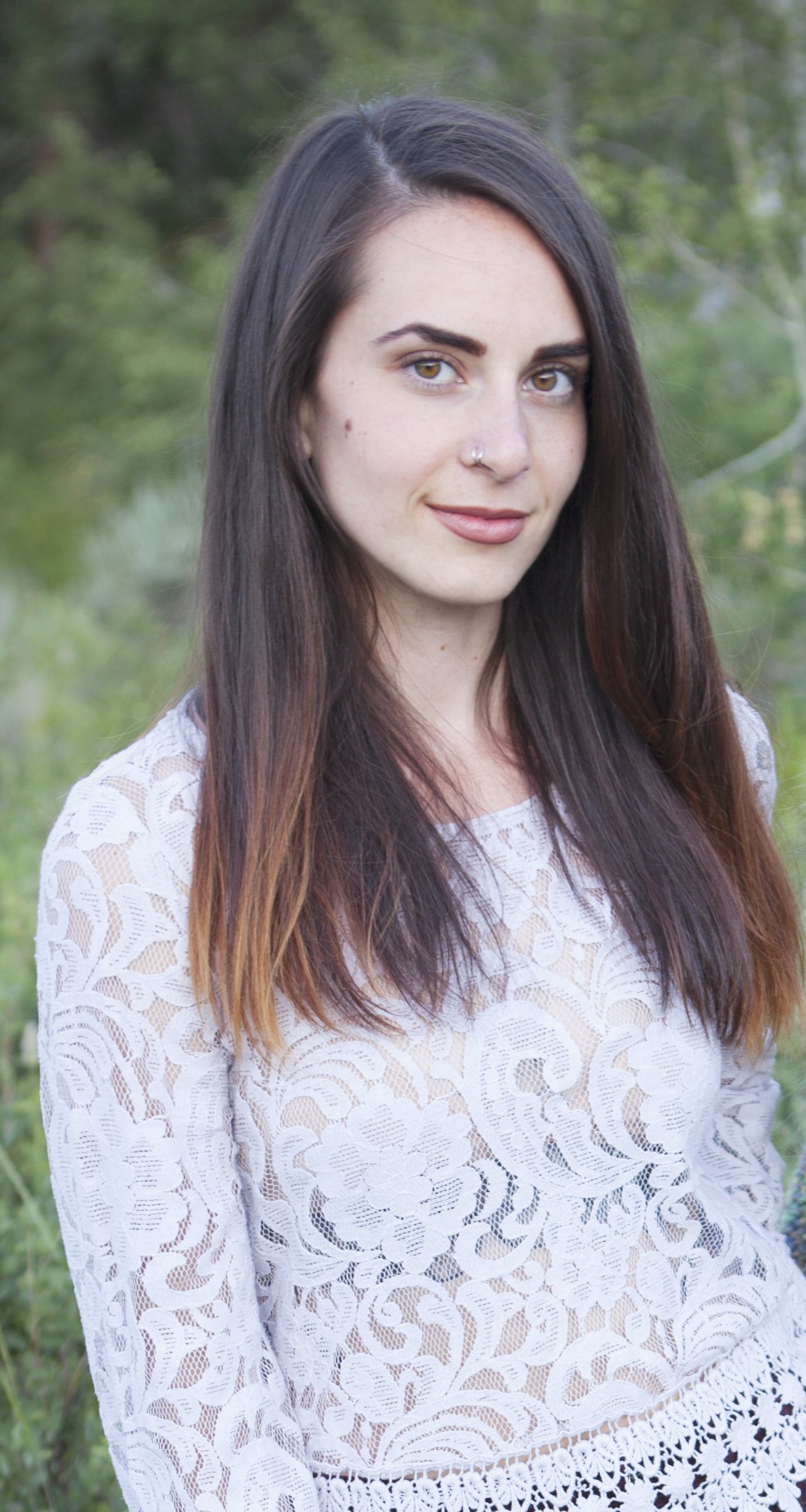 A young woman with long dark hair, light brown eyes, and a nose piercing, wearing a white lace top, standing outdoors in a green, leafy background.