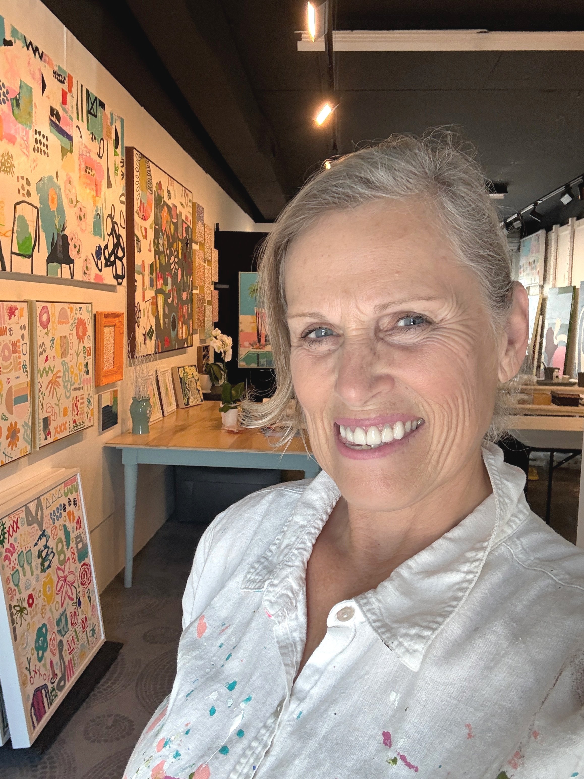 An abstract painting on a canvas with pink, green, white, and purple brushstrokes, hung on a white wall in an art studio with art supplies below.
