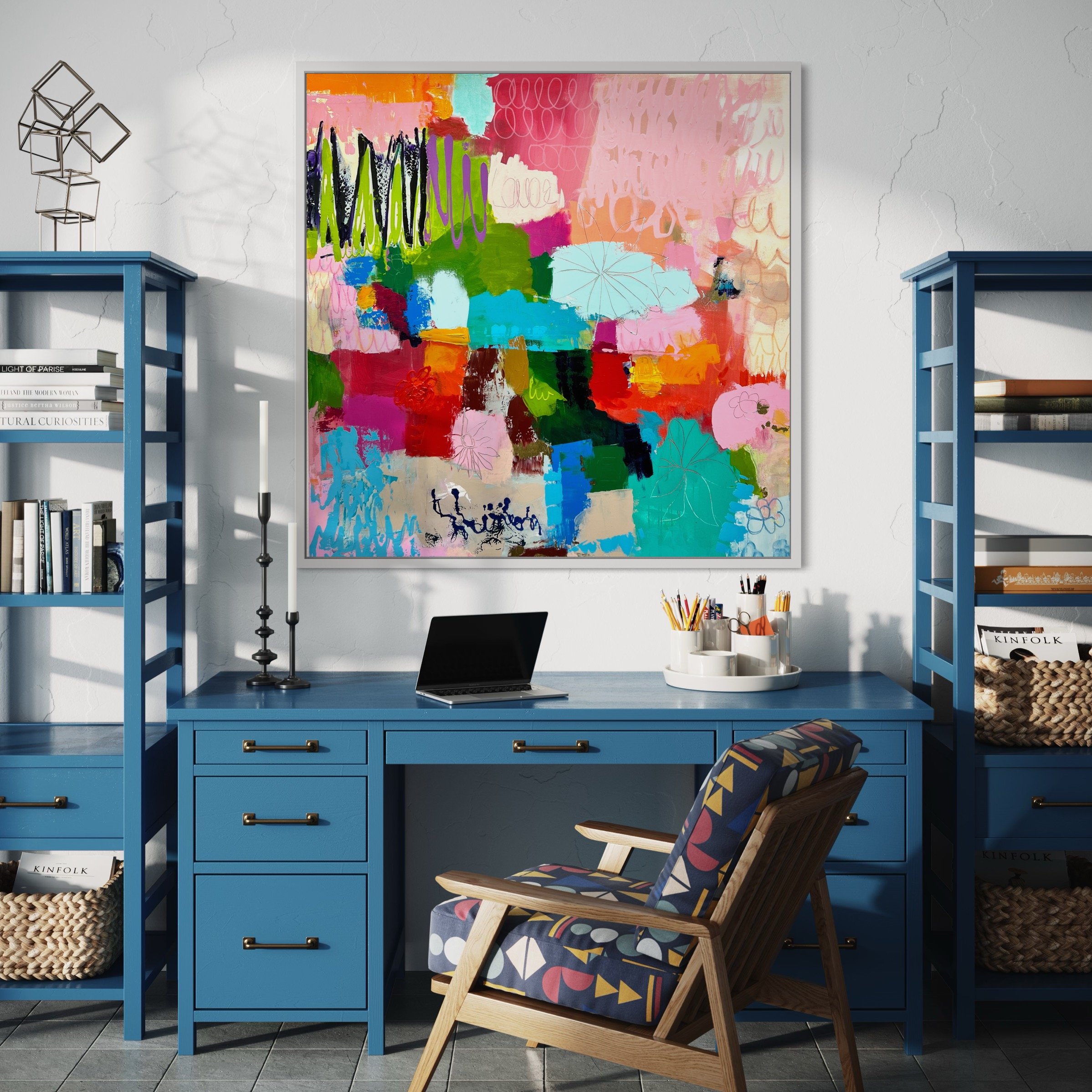 Interior view of a modern room with a large colorful abstract painting on the wall, a blue writing desk with a laptop, candle, and containers of pencils, a geometric patterned chair, and two blue bookshelves holding books and decorative items. The wa