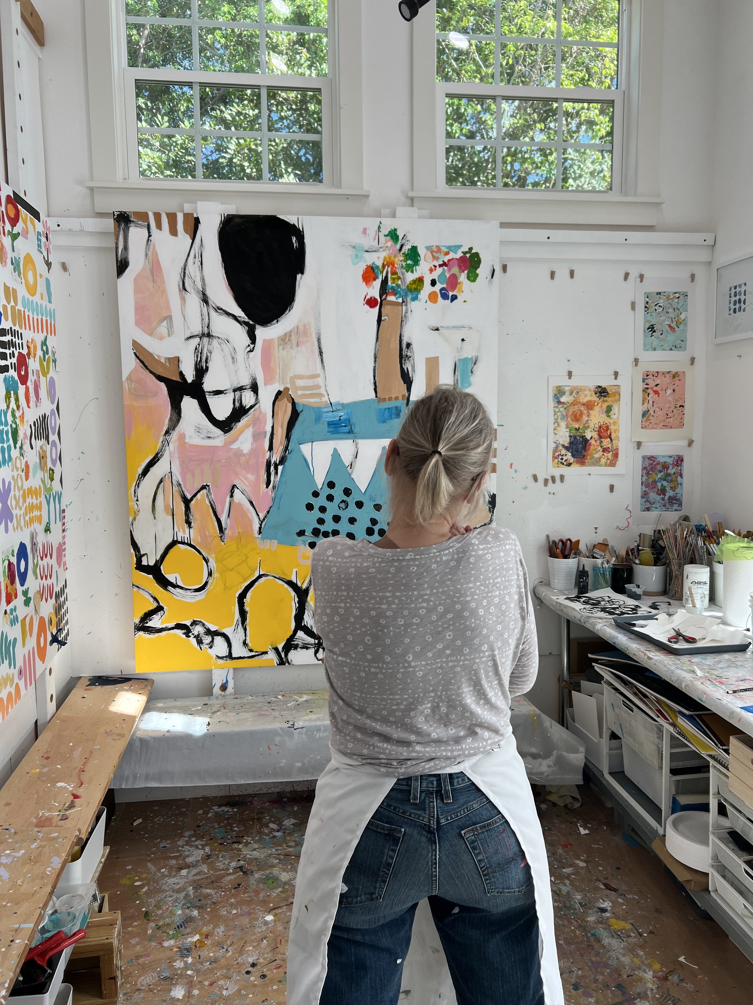 Multiple colorful abstract paintings displayed on a white gallery wall with large windows showing greenery outside.
