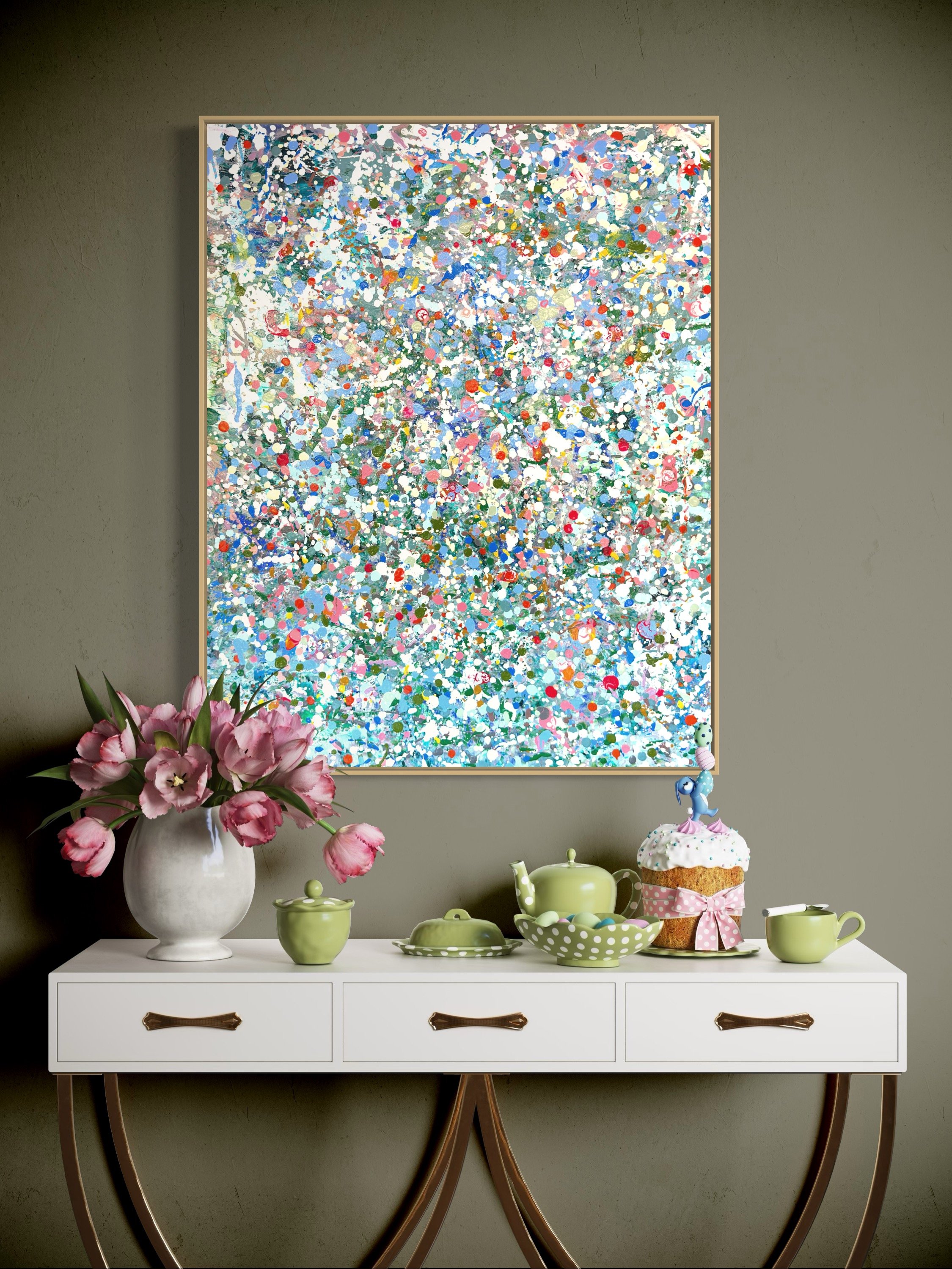 A white sideboard with brown handles. On top, a white vase with pink flowers, green cups, a bowl, a teapot, and a cake with colorful sprinkles and a pink bow. A large abstract colorful painting hangs on the wall behind.