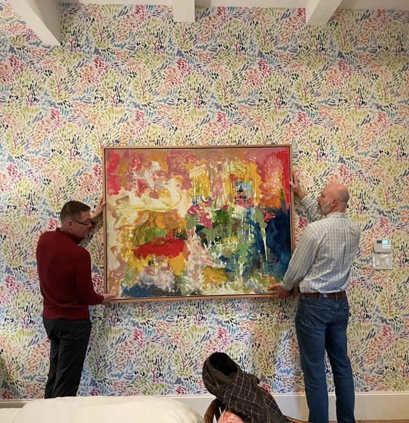 Two men are hanging a colorful abstract painting on a multicolored, patterned wall. One man is wearing a red sweater, the other a light plaid shirt. There is a person sitting on the floor in front of them, partially visible, with a brown and black ch
