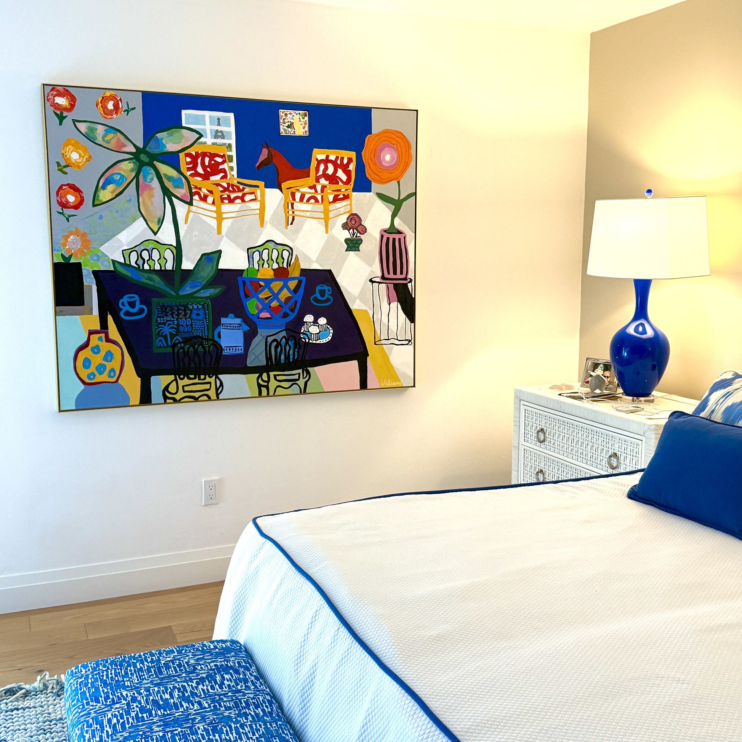A bedroom with three beds, a painting on the wall, a table lamp, and a framed photograph on a side table.