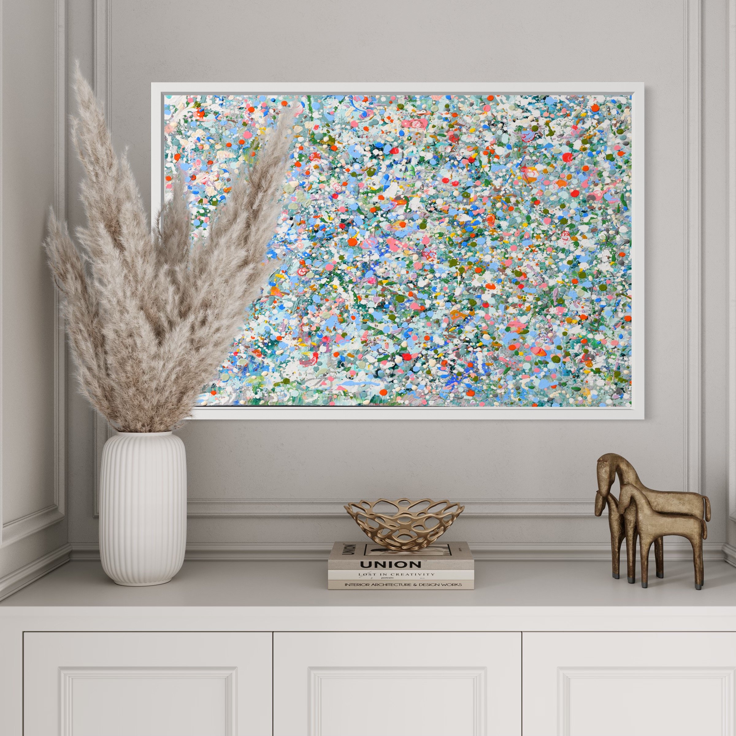 Interior of a room with a large colorful abstract painting on the wall, a tall white vase with pampas grass, a wooden decorative object, a closed white cabinet, and a stack of books titled 'UNION' and 'LOST IN CREATIVITY'.