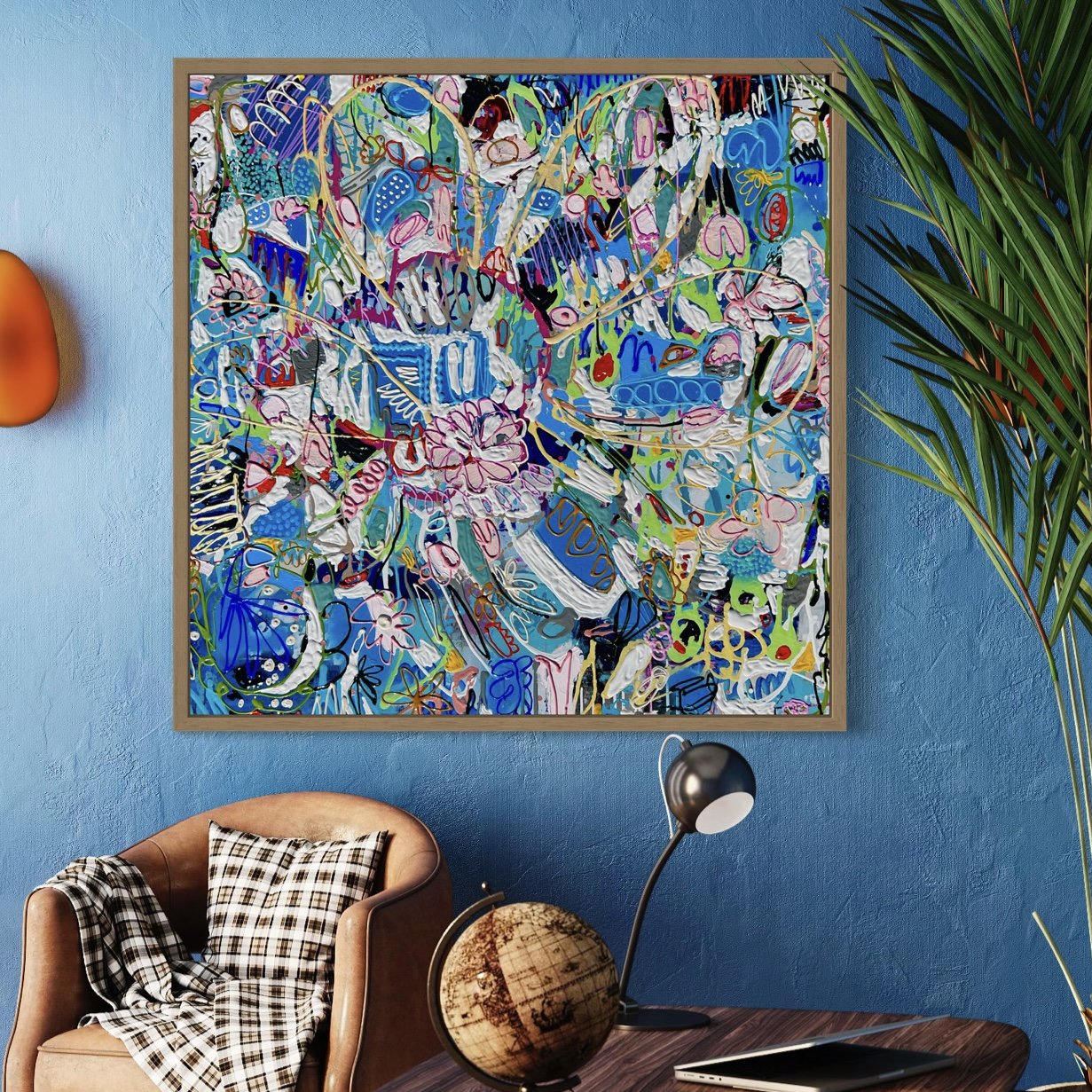 Colorful abstract artwork hanging on a blue wall in a modern living room, with a brown armchair, plaid blanket, globe, and desk lamp in the foreground, and a green plant on the right.