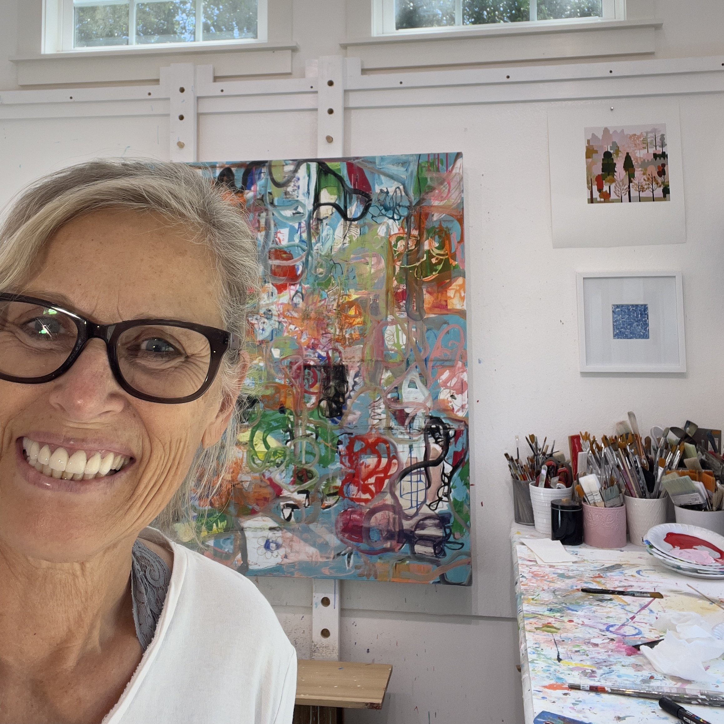 Room with three colorful abstract paintings on the walls, a wooden shelf below the paintings, and splatters of paint on the floor and on the paintings.