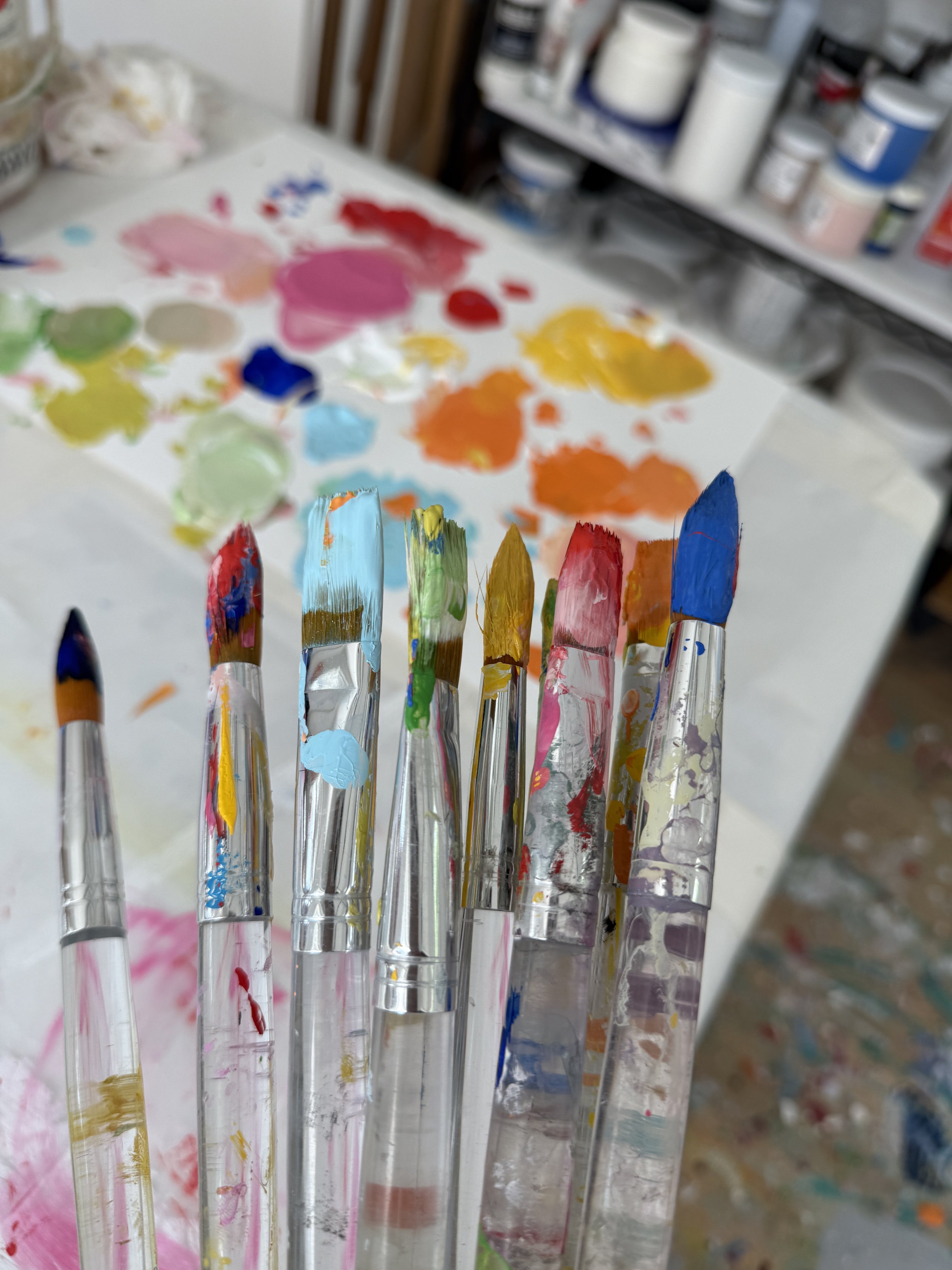 A wooden artist's workspace with many small containers of pastel and acrylic paint in various colors, next to abstract paintings with bright colors and bold brush strokes.