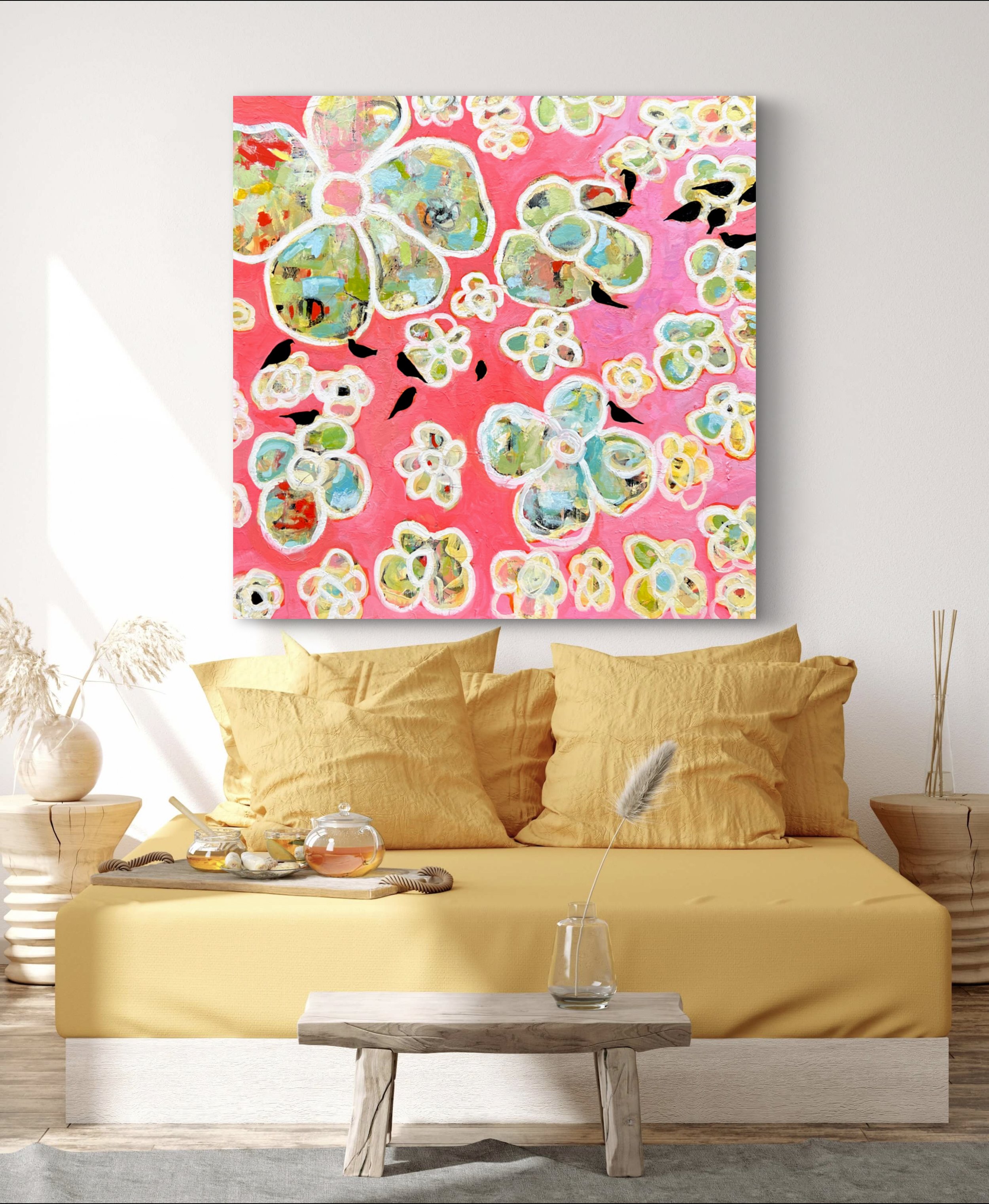 Colorful abstract painting hanging on a beige wall above a yellow sofa with large beige pillows, a white table with a wooden base in front, and small decor items including a glass of water with a feather. A small wooden side table with a white vase a