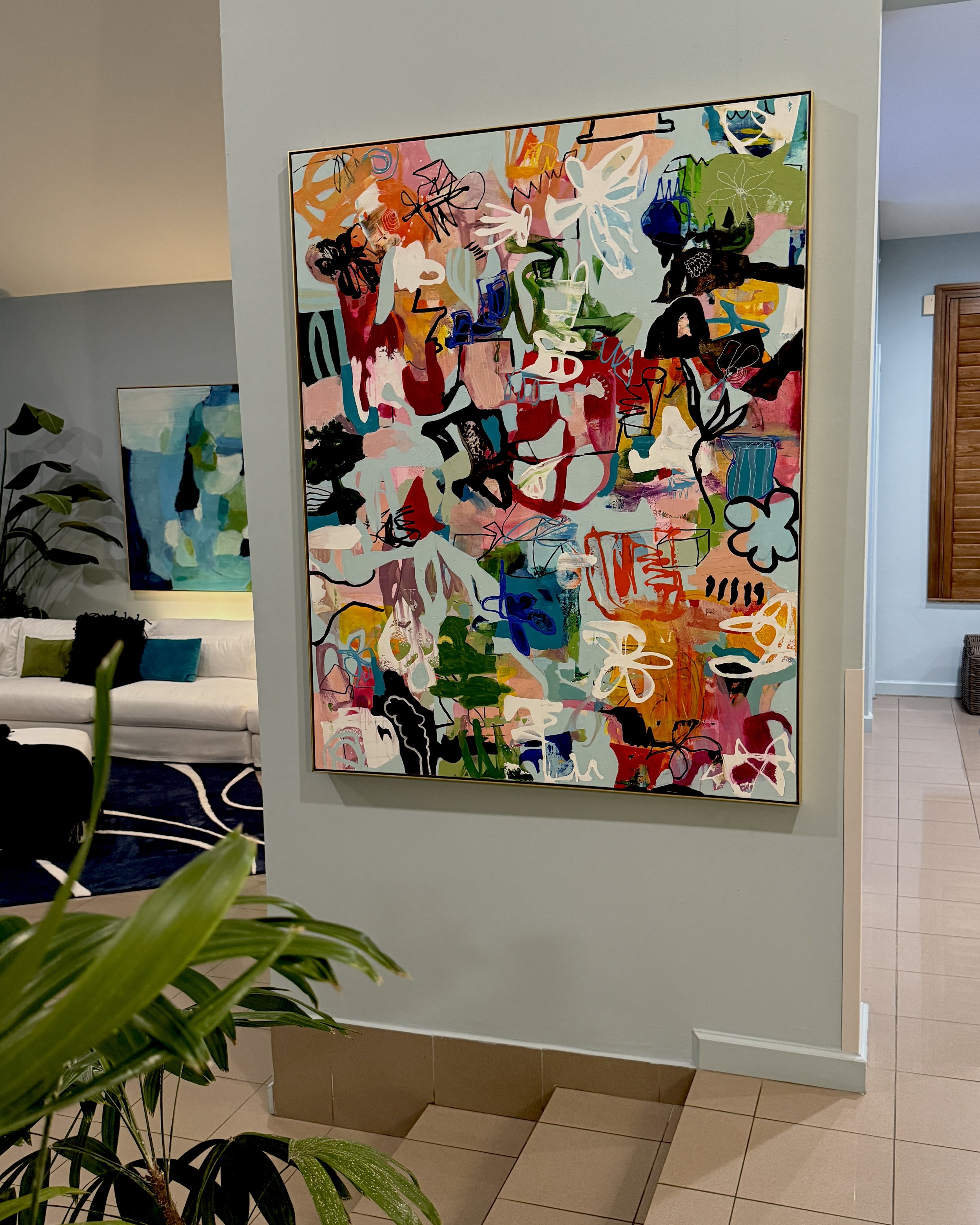 Colorful abstract painting on a wall in a room, with a white sofa and a green plant in the foreground.