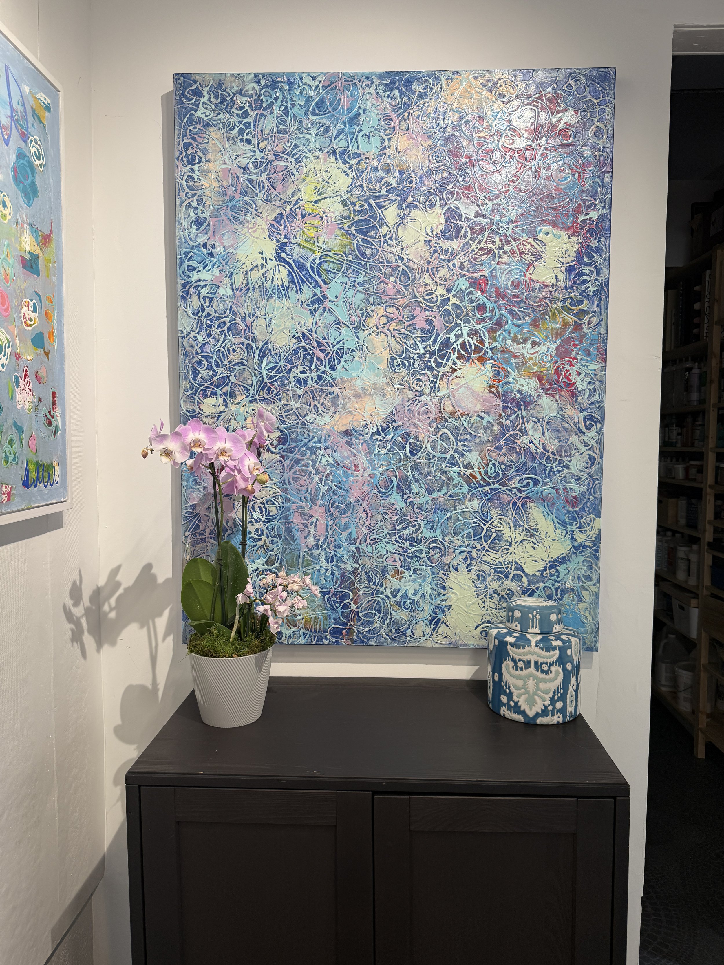 An arrangement of abstract artwork, a potted pink orchid, and blue patterned stools on a black console against a white wall in an art gallery.