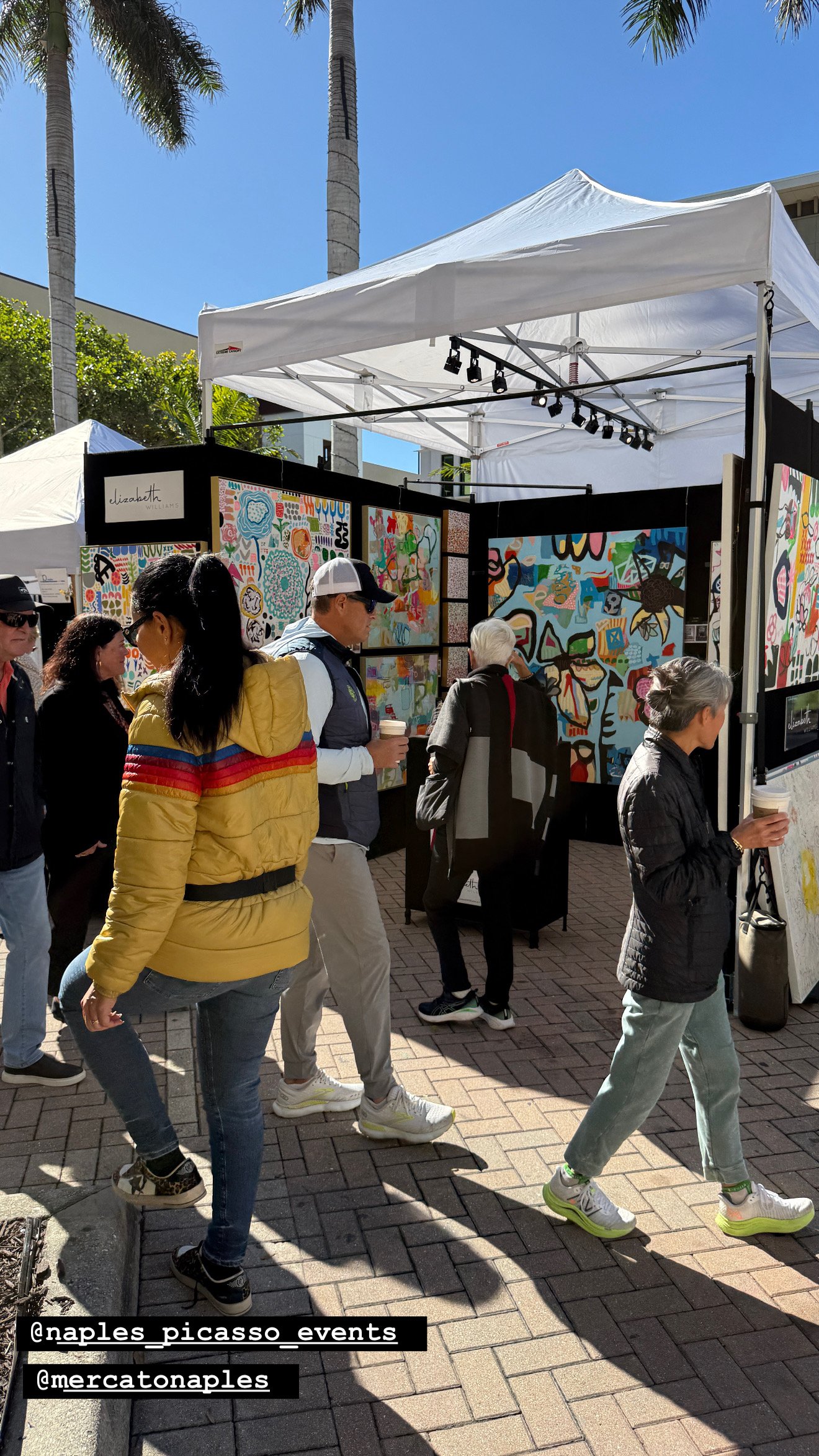 Visitors walking through an outdoor art festival booth viewing large colorful abstract paintings displayed on black walls under a white tent.