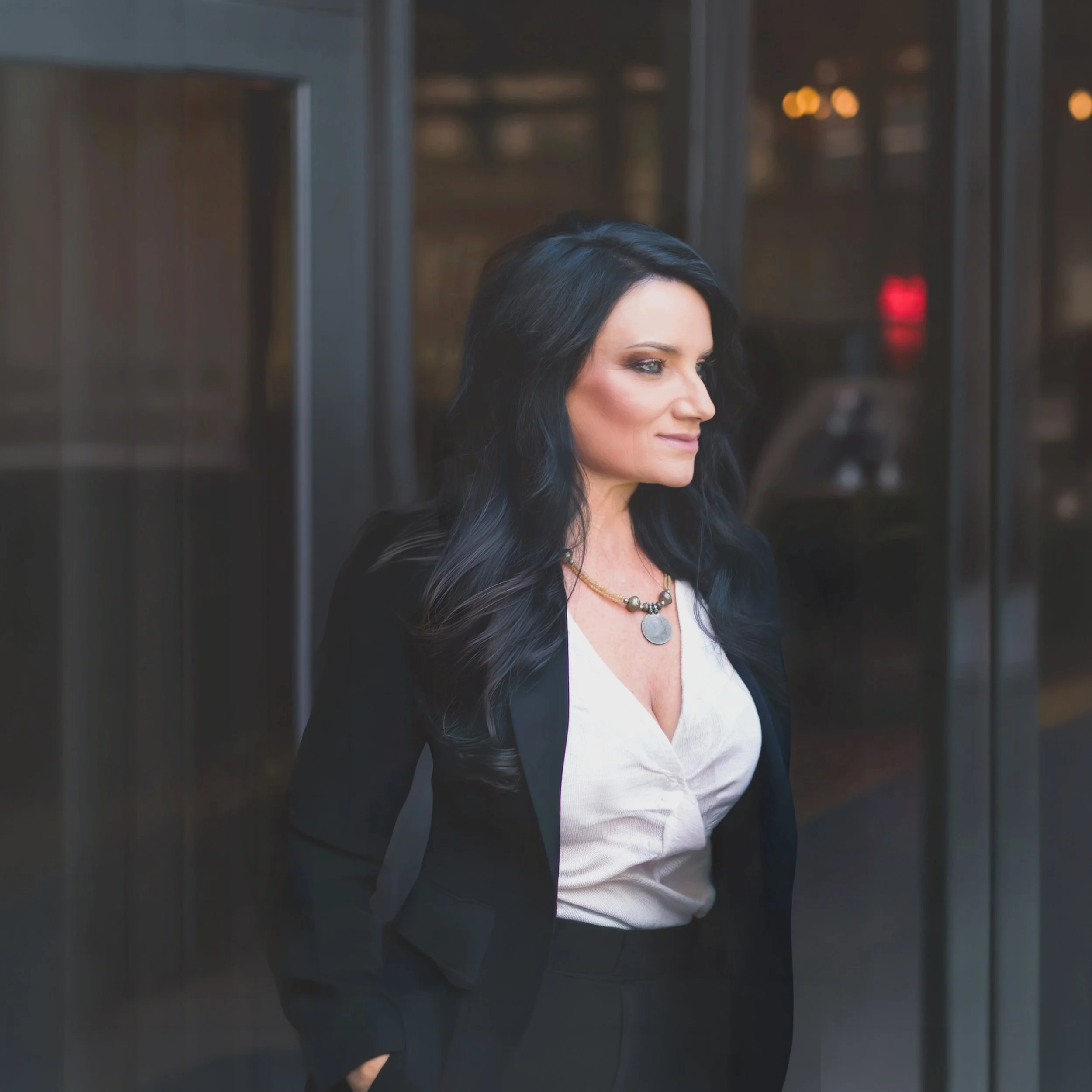 Sarah L. Stone, long dark hair wearing a white top and a black blazer, standing outdoors near a modern glass building with warm lighting.